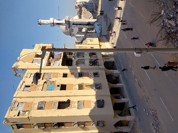 A street scene in Khan Yunis, Palestinian Territory, depicts widespread urban destruction under a clear blue sky. A multi-story, light-colored building on the left exhibits extensive structural damage, including numerous blast holes across its facade, missing windows, exposed interiors, and partial collapse of its upper levels and roofline. Arched architectural elements are visible on its ground and upper floors. A paved road with white lane markings, bisected by a streetlamp pole in the mid-ground, is flanked by dirt and piles of rubble. In the background, a white-domed mosque with a damaged minaret stands amidst further severely damaged or rubble-strewn structures.
Several individuals, including adults and children, occupy the street. In the foreground, two adults walk, while another stands further down the road. Children play on the pavement, with one riding a bicycle and another running. A small group of people congregates near the distant mosque. A light-colored vehicle is partially visible parked beneath the damaged building's ground-level archways. Strong shadows cast by the buildings and people indicate daytime. The scene reflects ongoing daily life amidst significant urban destruction.