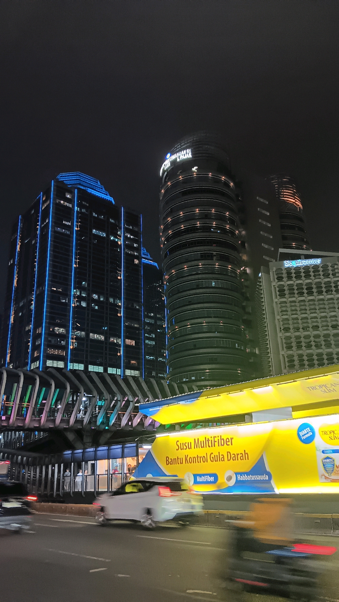 In the city of Central Jakarta, Indonesia, at night, a low-angle shot captures a vibrant urban scene. Dominating the background are several modern skyscrapers, with one building featuring prominent blue neon lights tracing its vertical lines and illuminated edges. To its right, a cylindrical building with tiered levels and horizontal lighting stands out. Further to the right, a blocky, light-colored building with a repetitive grid pattern of circular windows displays the signage "Squisecenter."
In the foreground, a bright yellow and blue billboard stretches across the frame, advertising "Susu MultiFiber Bantu Kontrol Gula Darah." The advertisement features images of fiber and what appears to be Habbatussauda (black seed). Below the billboard, a pedestrian overpass with a metallic structure and illuminated panels is visible.
On the street level, a white car is captured in motion, appearing as a blur due to motion. A motorcyclist, also blurred by movement, is visible further down the road. The street itself is asphalt, with visible lane markings. The overall atmosphere is that of a bustling city at night, with artificial lights from buildings and advertisements illuminating the dark sky.