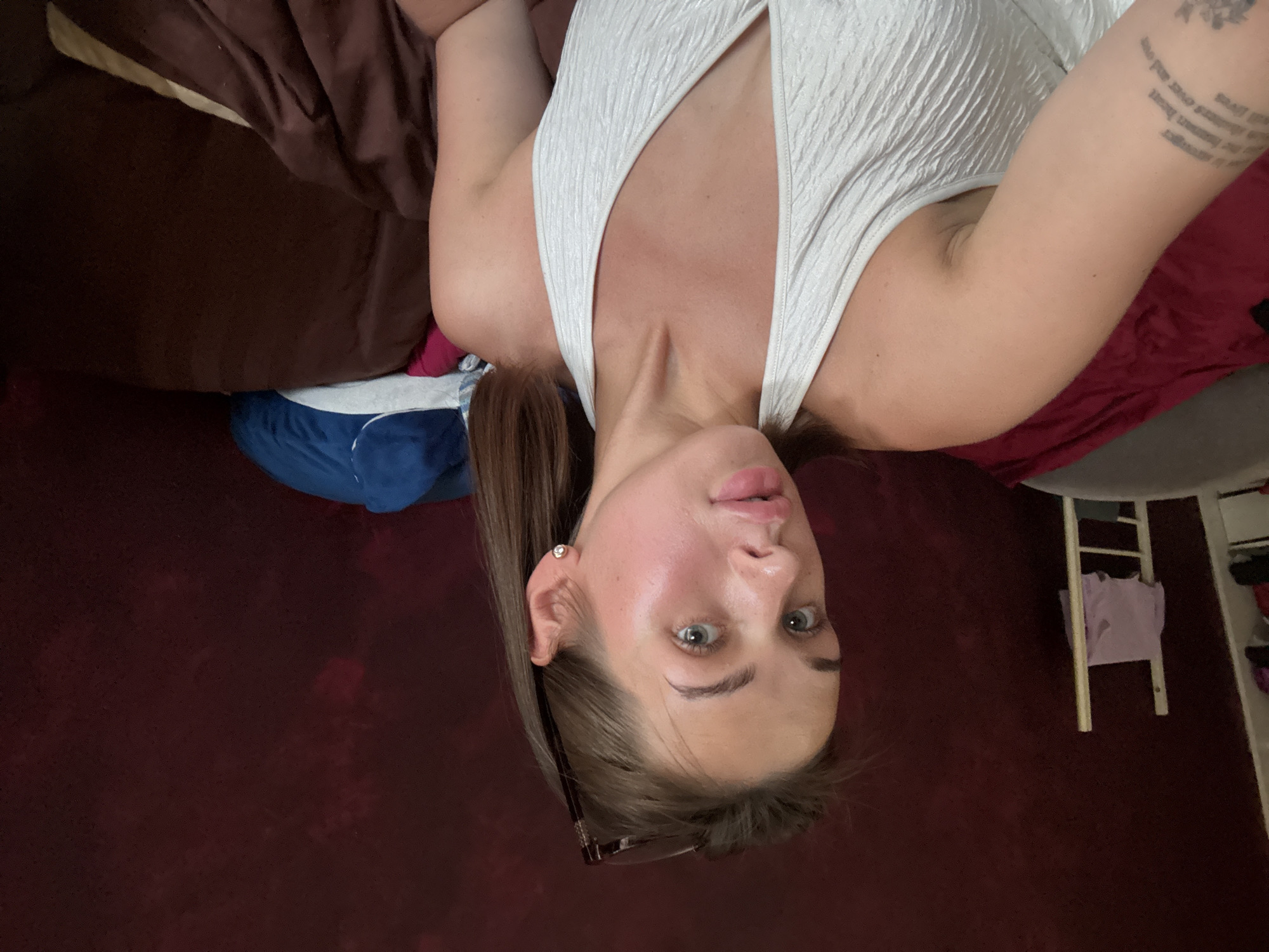 A young woman with light brown hair is taking a selfie in what appears to be a bedroom. She is wearing a white halter top. Her head is tilted to the left, and her eyes are looking towards the camera. She has sunglasses perched on top of her head. There is a tattoo on her left arm. The background shows a dark red wall and bedding in shades of brown and maroon. To the left of the woman, a white ladder with a pink garment draped over it is visible. The overall impression is of a casual, indoor setting. The image does not provide any specific clues to indicate the location in the city of Perm, Russia.