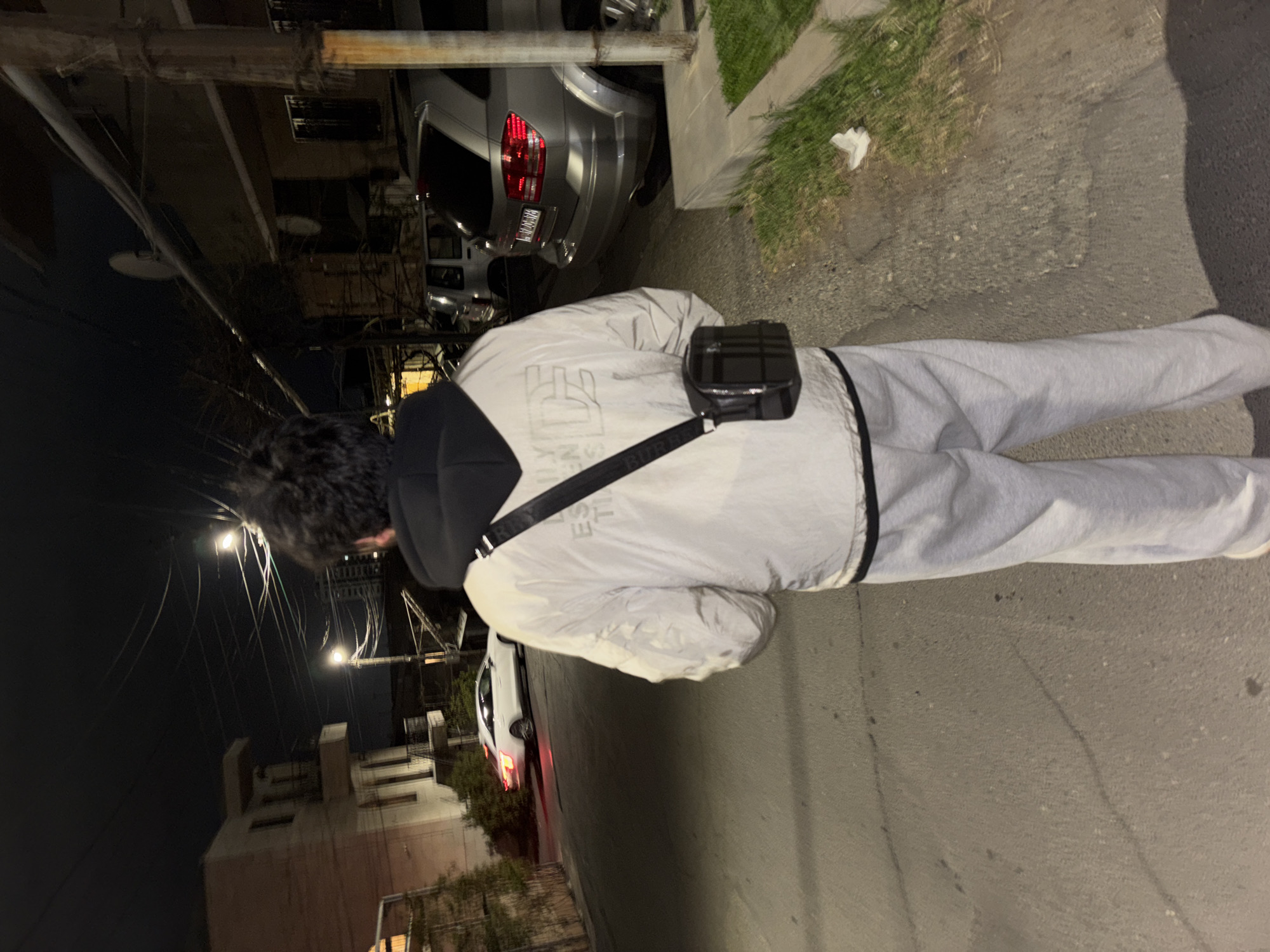 A person wearing a white Burberry jacket and gray sweatpants stands on a Yerevan street at night, seen from behind. The street is paved and leads past a parked silver SUV with a red taillight. Buildings line the left side of the street, with streetlights illuminating the scene. Green foliage is visible to the right.