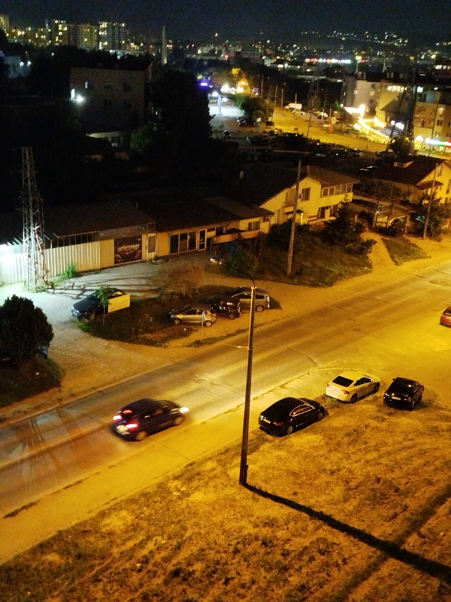 The image is an aerial night view of a street scene in Varna, Bulgaria. A two-lane road runs diagonally across the frame, with cars parked on the right side and one in motion on the left side. Adjacent to the road is a grassy area and beyond the road a building with a sign in Cyrillic that reads "АВТОСЕРВИЗ" (Auto Repair). There are parked cars in front of the auto shop. In the background, a residential area is illuminated by street lights. The overall lighting suggests it is nighttime, with sodium street lights casting an orange hue.