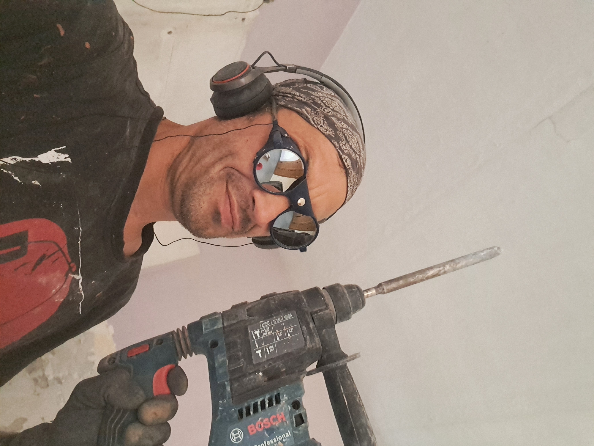 The image features a man in Macello, Italy, wearing a bandana, headphones, and safety goggles, holding a blue and black Bosch professional rotary hammer drill. He is wearing a black t-shirt with red paint splatters and a dark glove on his left hand. The man is looking down at the drill, and the camera is positioned below him, looking up. The background is a pale, textured wall, possibly an interior wall under construction or renovation, with some light fixtures or wires visible on the ceiling. The lighting suggests it is daytime, possibly indoors. The man appears focused on his task, with a slight expression of concentration. The drill has visible markings, including the "Bosch" logo and "Professional." The goggles reflect the surroundings, showing a blurred image of a room.