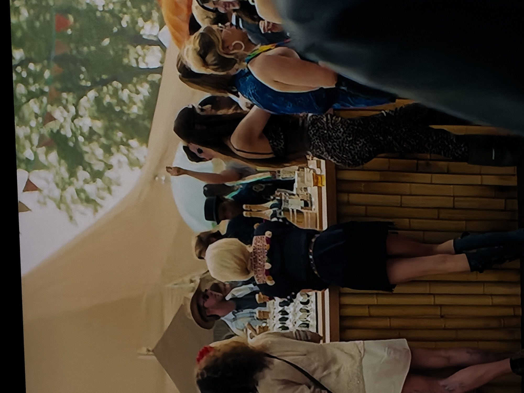 A vibrant outdoor gathering is depicted, with several individuals congregated around a temporary bar or serving station. The image is oriented with the people standing sideways, so for this description, they are considered upright.
In the foreground, a person with blonde hair wearing a blue top and large hoop earrings stands, possibly engaging with others or observing the display. Next to them, a person with dark hair is seen from the back, dressed in a dark, possibly animal print, top and trousers.
Further along the bar, a person with blonde, short hair and a dark top adorned with colorful embellishments stands, looking towards the bottles. Behind the bar, a person in a hat and a light-colored shirt is visible, likely serving or attending to the patrons.
The bar itself is constructed from bamboo-like material, and shelves behind it are stocked with numerous bottles, mostly clear, containing golden or amber liquids, suggestive of alcoholic beverages. Various glasses are also arranged on these shelves.
In the background, a large white tent or canopy is visible, suggesting an outdoor event. Lush green foliage and trees indicate a natural setting, and a few small red triangular flags are faintly visible, possibly part of decorative bunting. A large white balloon or similar round object floats near the bar area.
The scene appears to be a social event, perhaps a tasting or a casual celebration, taking place during the daytime in good weather. People seem relaxed and engaged in the activity. No visible text is present in the image. The location context provided is Sofiivska Borshchahivka, Ukraine.