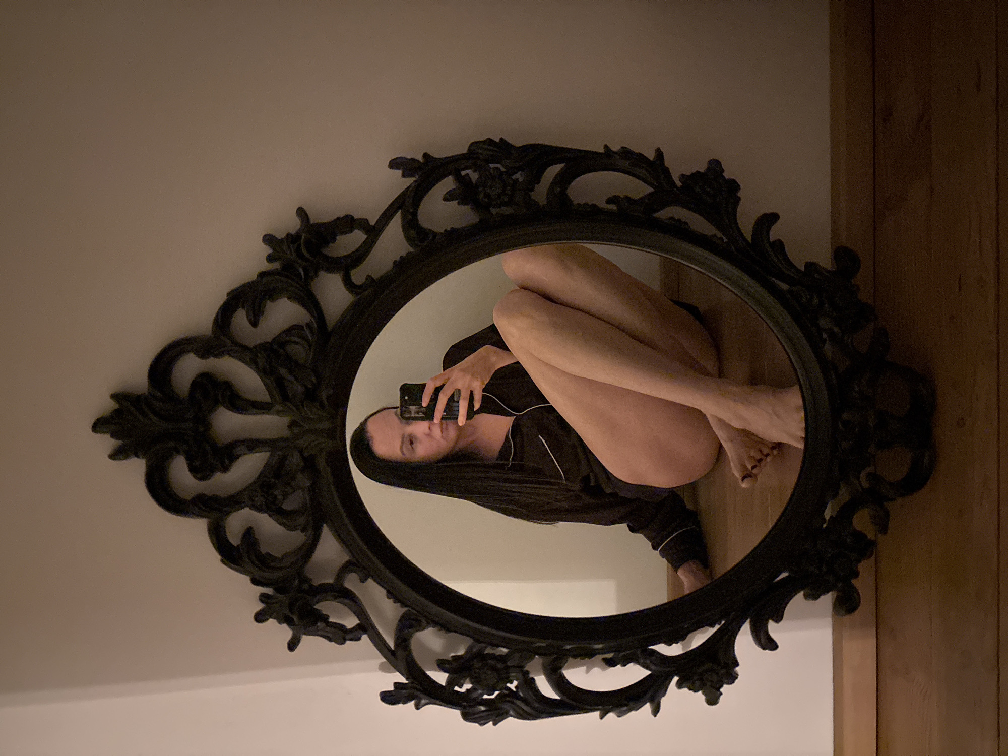 The image shows a woman reflected in an oval, ornate black mirror. She is holding a cell phone up to take a picture of herself in the mirror. She is wearing a dark-colored pajama set with white piping. She is sitting on a floor that appears to be wooden, with the wall behind the mirror painted a light color. The location is unknown and not related to Chaikovsky, Russia.