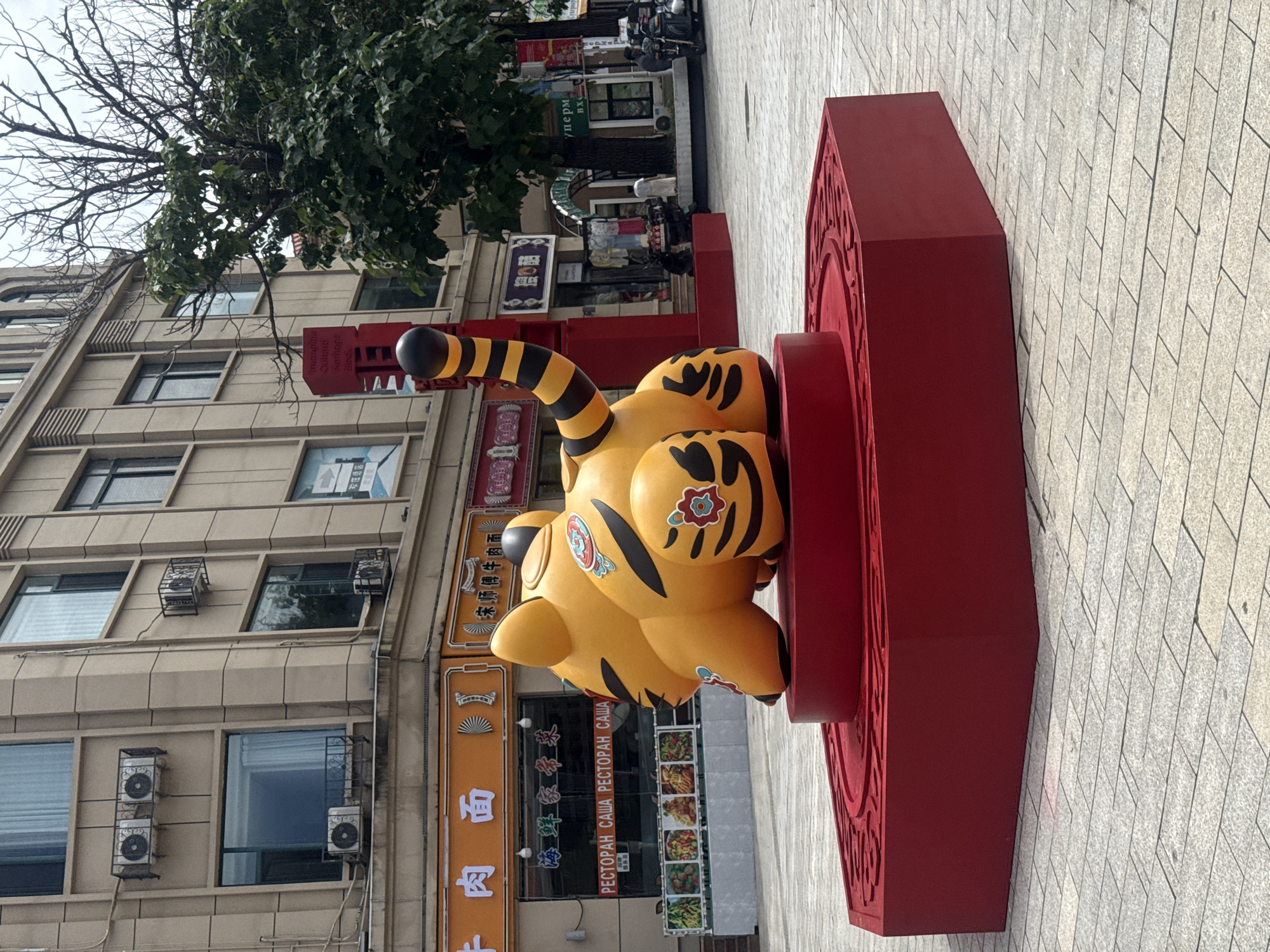 This is a medium shot of a large, stylized tiger sculpture in a city square. The sculpture is bright yellow with black stripes and decorative floral patterns on its body. It is resting on a hexagonal red pedestal. The background features a multi-story building with several windows and air conditioning units. Below the building, there are storefronts with Chinese and Russian signage. One storefront appears to be a restaurant with images of food displayed. A red pillar with black and yellow stripes stands to the right of the tiger sculpture. In the distance, there are trees, other buildings, and a few people, as well as a motorcycle. The ground is paved with light-colored rectangular stones. The image was taken in Qinhuangdao, China, likely during daytime.