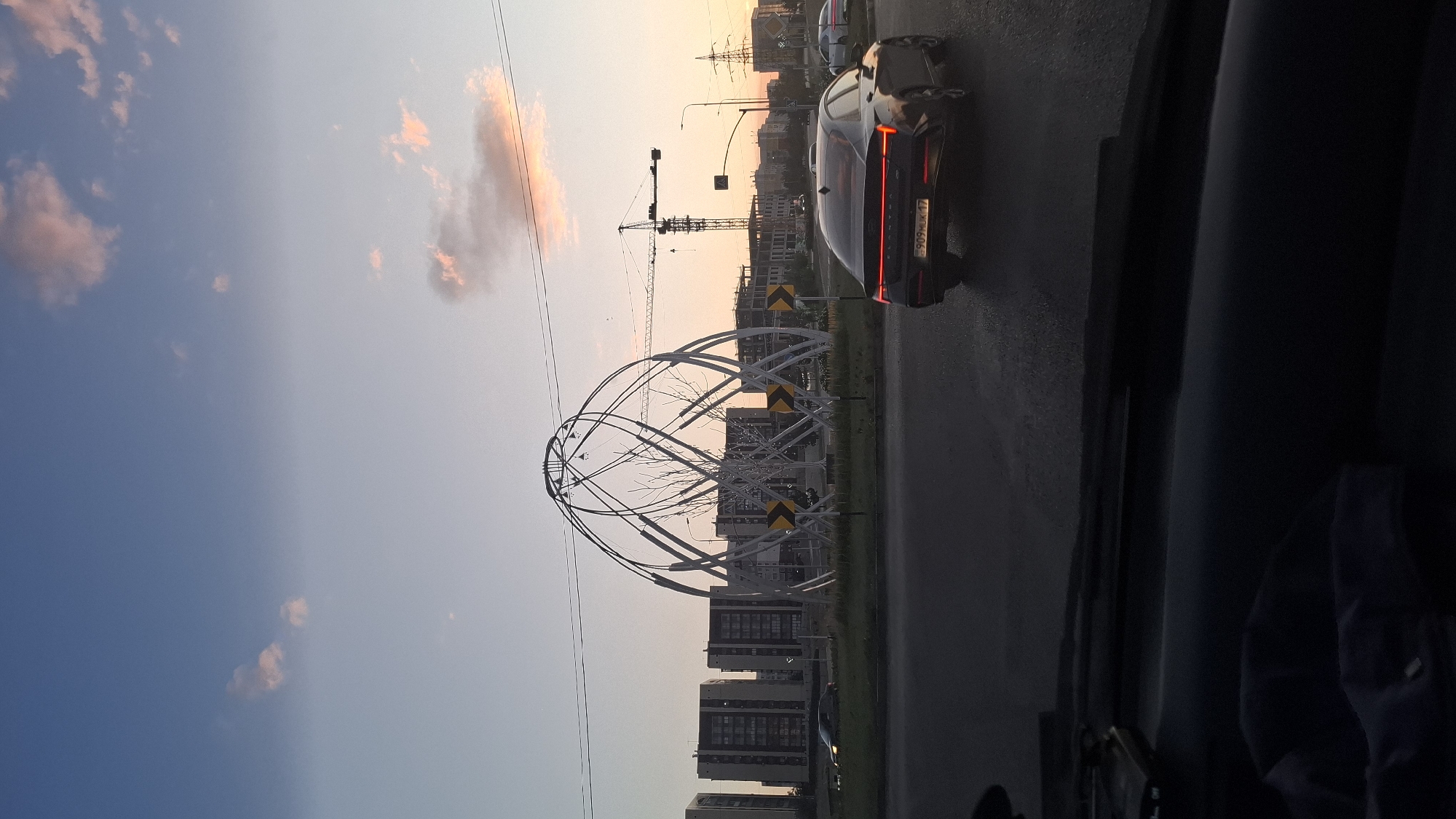The image depicts an urban scene at dusk, likely in Shymkent, Kazakhstan, centered around a large, modern sculptural installation. The sculpture, with its intricate white, curved metal framework, dominates the middle ground, appearing to represent abstract, flowing shapes or perhaps stylized plant-like forms. Behind the sculpture, several multi-story residential buildings are visible, characterized by their uniform architecture and glass-paneled facades, suggesting a contemporary urban development. Construction cranes and unfinished buildings to the right indicate ongoing development in the area.
The foreground is occupied by a dark car's interior, suggesting the photo was taken from a vehicle. A black sedan is prominently featured on the wet asphalt road, its taillights illuminated, indicating it is moving or stopped in traffic. The license plate of this car is partially visible, showing Cyrillic characters and numerals. The road surface appears wet, reflecting the ambient light and suggesting recent rain or a damp environment.
The sky above is a gradient of soft blue and pale orange, indicative of sunset or sunrise, with scattered clouds tinged with pink. Power lines and streetlights are also discernible, typical elements of an urban landscape. The overall impression is that of a modern city at the end of the day, with functional infrastructure, residential areas, and public art blending into the evening light. The presence of the sculpture and the architectural style of the buildings suggest a location that is likely a public square or a prominent roundabout designed as an urban landmark.