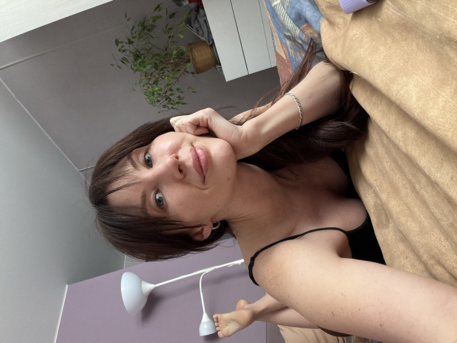 The image depicts a single adult female in a domestic bedroom setting, captured in a selfie-style portrait. The subject is lying prone on a bed, facing the camera with a slight smile and direct eye contact. She has long, dark brown hair with bangs and appears to be in her 20s to 30s. She is wearing a black spaghetti-strap top, small silver hoop earrings, and a silver-colored braided bracelet on her left wrist. Her left hand supports her chin. Her bare feet are visible, elevated against a wall.
The bed is covered with a light brown blanket, partially revealing a multi-colored patterned sheet or duvet with blue, brown, and muted red/purple tones. The background includes a light purple wall to the left and a light gray wall to the right. A white floor lamp with two light heads is positioned on the left side of the frame. On the right side, a potted Ficus plant in a woven brown basket sits atop a white dresser or shelf, with some folded textiles visible beside it. The lighting is even, suggesting natural illumination. The specific location within the city of Perm, Russia, is not discernible from the visual information provided.