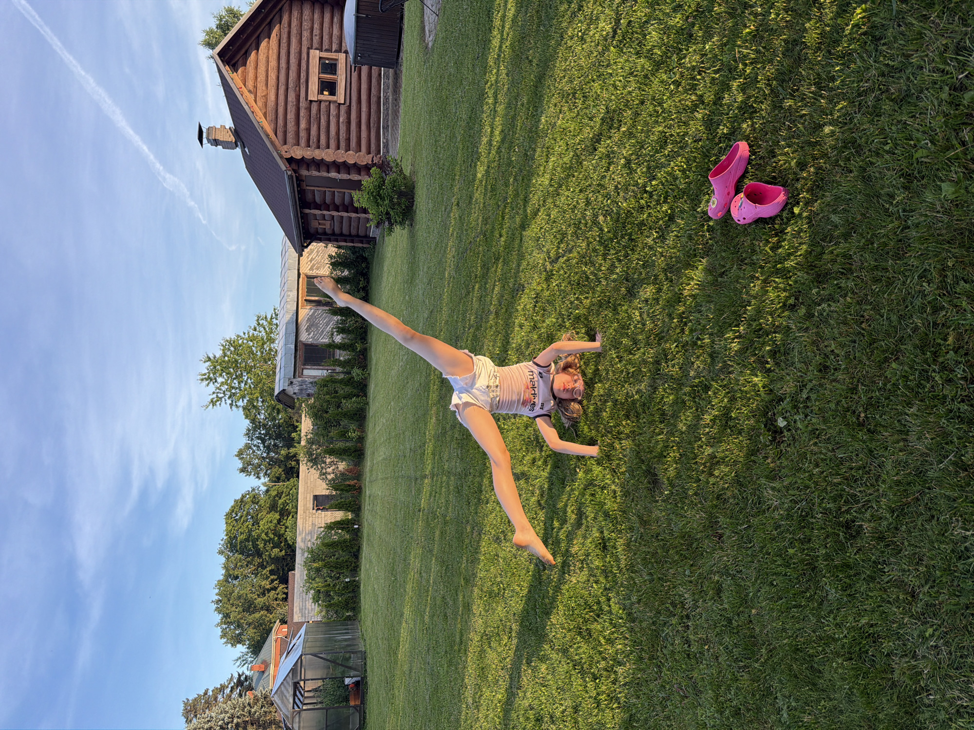 A young girl is lying on her back on a grassy lawn, with her legs spread apart and her right leg extended upwards in a split. She is wearing white shorts and a light-colored t-shirt with some text on it. Her eyes are closed, and she has a slight smile on her face, suggesting she is enjoying herself. She is wearing sunglasses. Next to her on the grass are a pair of bright pink clogs.
The scene appears to be a backyard, with a wooden cabin-like structure in the background. There is also a greenhouse visible, as well as trees and other vegetation. The sun is shining, casting long shadows across the grass, indicating it is likely late afternoon or early evening. The sky is blue with some wispy white clouds. The overall impression is one of a relaxed and playful summer day.
The text on the girl's t-shirt is partially visible and appears to say "matrìes" with some other unclear lettering.
