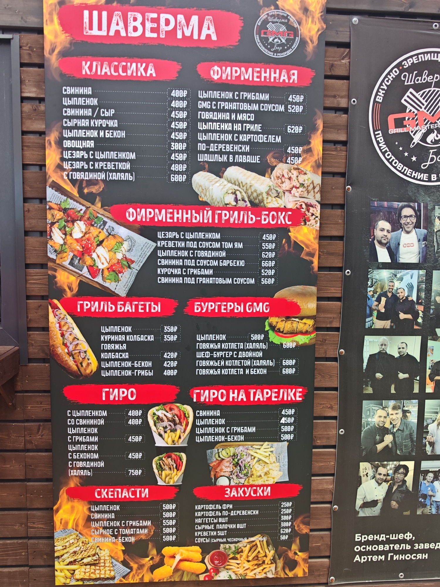 The image shows a food menu board with various options like "Shaverma," "Classic," "Signature Grill-Box," "Grill Baguettes," "Burgers GMG," "Gyros," and "Snacks." The menu is in Russian and displays prices in rubles. There are also pictures of food items like wraps, burgers, and fries. The background features graphics of flames. On the right side of the board, there are several photographs of people, some posing together and others individually. The text "Brand Chef, Founder Artem Ginosyan" is visible at the bottom right. The menu board is mounted on a wooden surface, suggesting it is an outdoor establishment. The presence of Russian text and a Russian restaurant name implies a location in Russia, possibly a resort town given the casual presentation of the menu. While no explicit geolocation data is available, the mention of "Sukko, Russia" in the prompt suggests this is a food stall or cafe in Sukko, Russia.