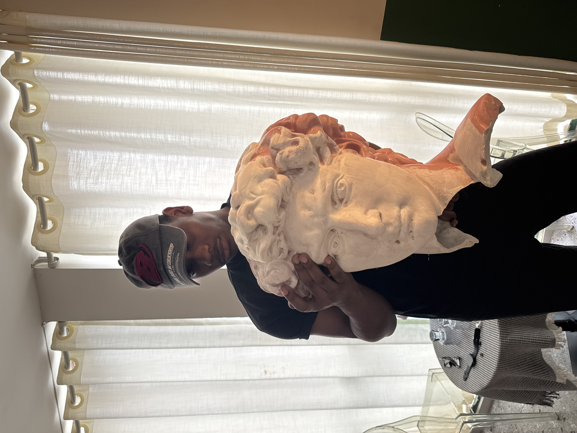 In an indoor setting in Liangjiang, China, a male individual is positioned centrally, holding a fragmented plaster bust. The individual, appearing to be of Black ethnicity, wears a dark grey baseball cap with a red and white logo, a black short-sleeved t-shirt, and black pants. A ring is visible on his left ring finger. He looks downward towards the bust, which he supports with both hands, positioned around its upper chest and neck.
The bust, depicting what appears to be the head and upper shoulders of Michelangelo's David, is predominantly white plaster with exposed reddish-orange material, possibly terracotta or clay, visible in areas such as the hair and fragmented neck. The bust displays detailed facial features and curly hair.
To the left of the individual, a small round table covered with a grey and white checkered tablecloth featuring white fringe is visible. On the table are two clear glasses, a black object resembling a smartwatch, and a pair of eyeglasses. A transparent, clear chair is partially visible behind the table. The background features floor-to-ceiling white sheer curtains with grommets, suspended from a light-colored rod. To the right, a plain light-colored wall transitions to a dark green section closer to the floor. The room is brightly lit, with light originating from behind the curtains.