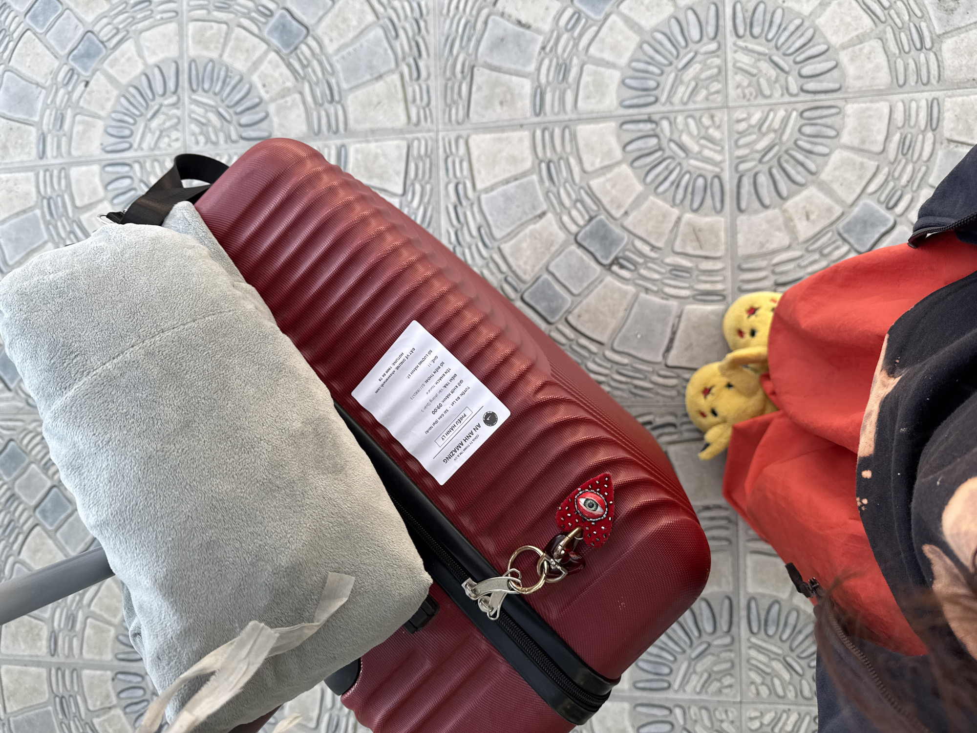 An overhead shot shows travel luggage on a patterned tiled floor, likely in Da Lat, Vietnam, given the context. A red, ribbed suitcase is prominently displayed, topped with a gray, plush blanket. A luggage tag is affixed to the suitcase, bearing Vietnamese text. Attached to the suitcase zipper is a keychain with a heart-shaped charm featuring an eye. To the lower right of the frame, partially visible, are a pair of yellow plush slippers and a red bag. The ground appears to be an outdoor paved area with decorative tile patterns.