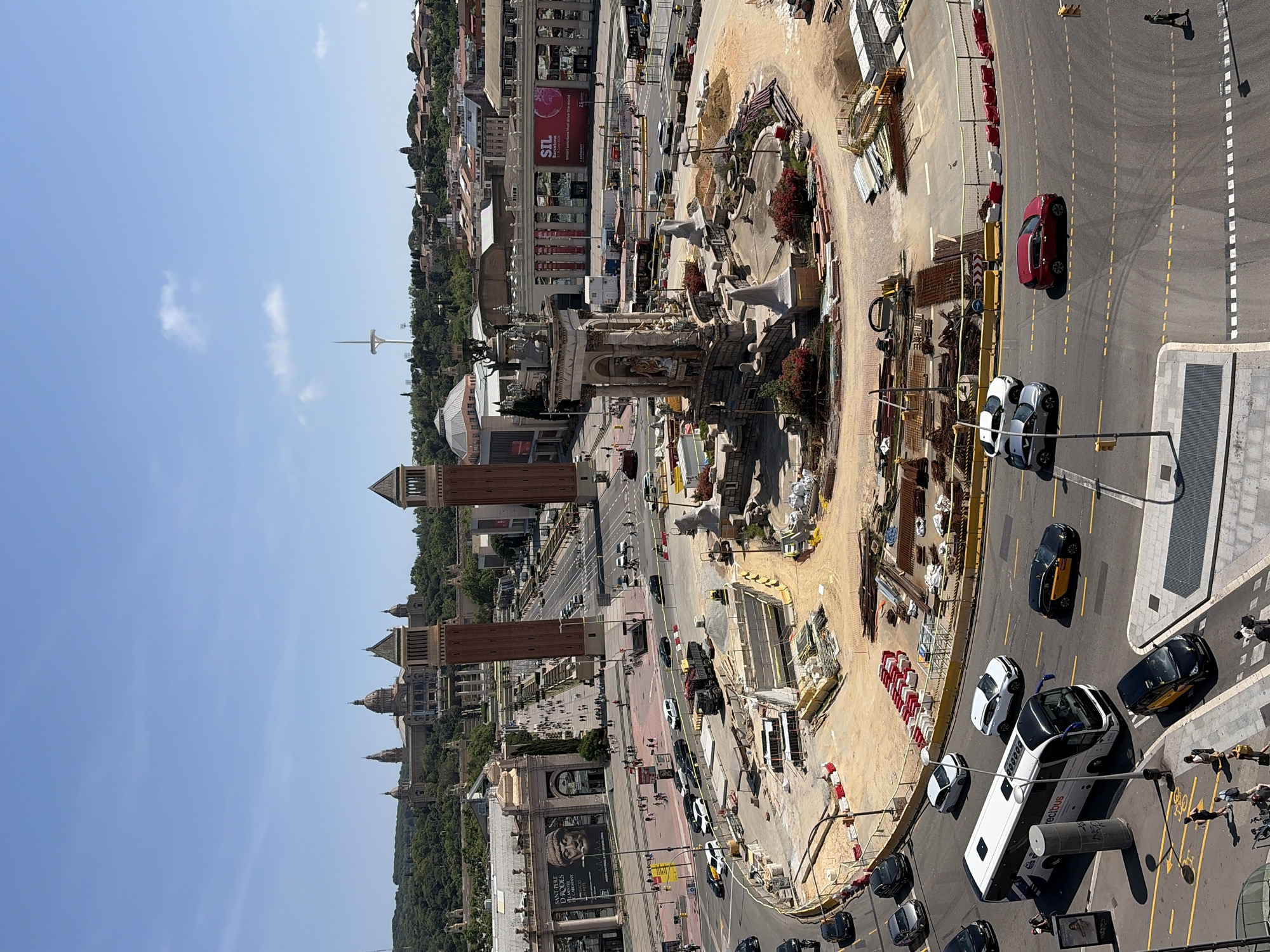 The image shows an aerial view of Plaça d'Espanya in Barcelona, Spain, undergoing construction. The Magic Fountain of Montjuïc is visible in the center of the plaza, surrounded by construction equipment and materials. The Venetian Towers and the National Art Museum of Catalonia (MNAC) are visible in the background. Cars and buses circulate around the plaza. Pedestrians are walking on the sidewalks. The environment is urban, with clear blue skies. A tall radio tower is visible in the background.