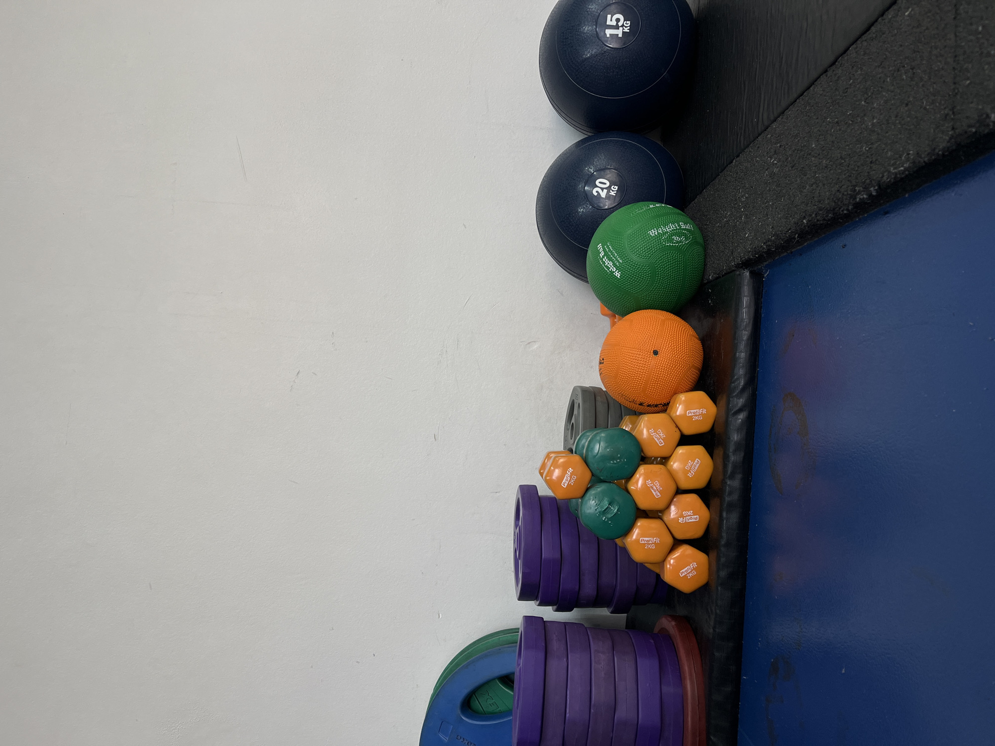 This image captures a collection of fitness equipment neatly organized in what appears to be a gym or training facility in Krasnoyarsk, Russia. The scene features a clean white wall on the left, transitioning to a dark blue floor mat or covering on the right, separated by a thick black textured strip.
Various pieces of exercise equipment are visible, arranged along the white wall and black strip. These include several medicine balls: two large, dark blue balls labeled "15 KG" and "20 KG" respectively, a smaller green ball marked "Weight Ball 2KG" and "Profi Fit", and an orange ball. Below these, a cluster of small, hexagonal dumbbells is stacked, predominantly orange with a few green ones mixed in. Each of these dumbbells is labeled "Profi Fit 2KG". Further down, several purple weight plates are stacked horizontally, likely for barbells. At the very bottom left, parts of larger green and blue weight plates are visible, with the text "LEX" discernible on the blue one.
No people are present, and no activity is taking place; the equipment is stored and ready for use. The lighting suggests an indoor setting, but the time of day and weather are not discernible. The vibrant colors of the fitness gear contrast with the neutral white wall and dark blue floor, creating an organized and functional environment.