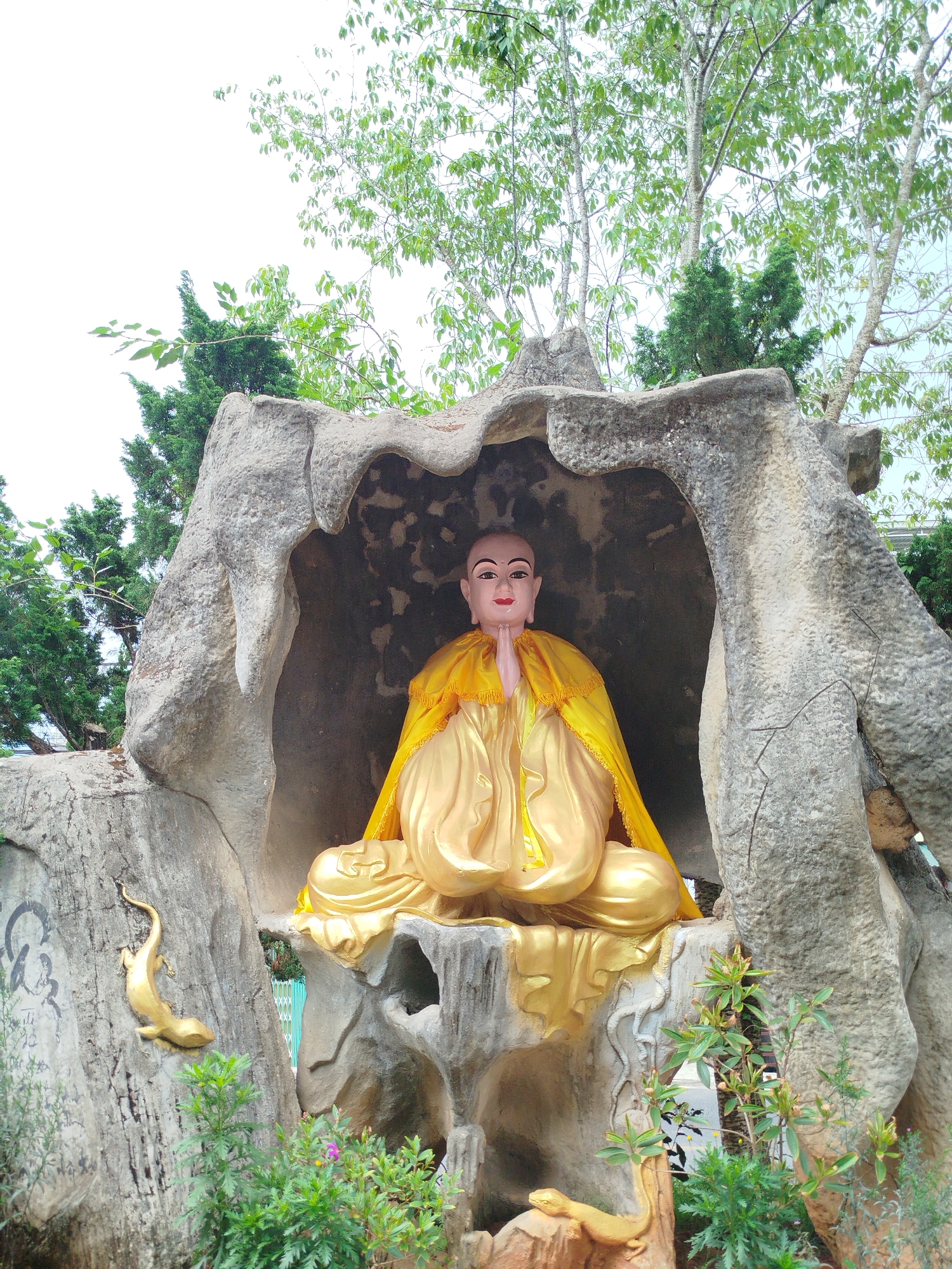 A serene statue of a Buddhist monk or spiritual figure is depicted seated in a meditative posture within a man-made rock grotto. The figure, centrally positioned and facing forward, has a bald head, pale skin with reddish facial accents, and a tranquil expression. It wears flowing, bright golden robes, with hands clasped in the Anjali Mudra.
The grotto is constructed from a grey, textured material resembling natural rock, providing a cave-like enclosure for the statue. On the left side of the rock formation, two golden lizard or gecko sculptures are visible, one partially obscured by lush green plants at the base, and another prominently displayed higher up. Faint, dark, illegible markings or graffiti can be seen on the left side of the rock structure.
The outdoor setting is characterized by abundant green foliage and tall trees with light-colored trunks, reaching upwards towards a bright, overcast sky, indicating daytime. Various small plants and shrubs, some bearing tiny blossoms, grow around the base of the rock formation. The scene evokes a sense of peace and contemplation, typical of a spiritual garden or temple in Cam Ly - Đà Lạt, Vietnam.