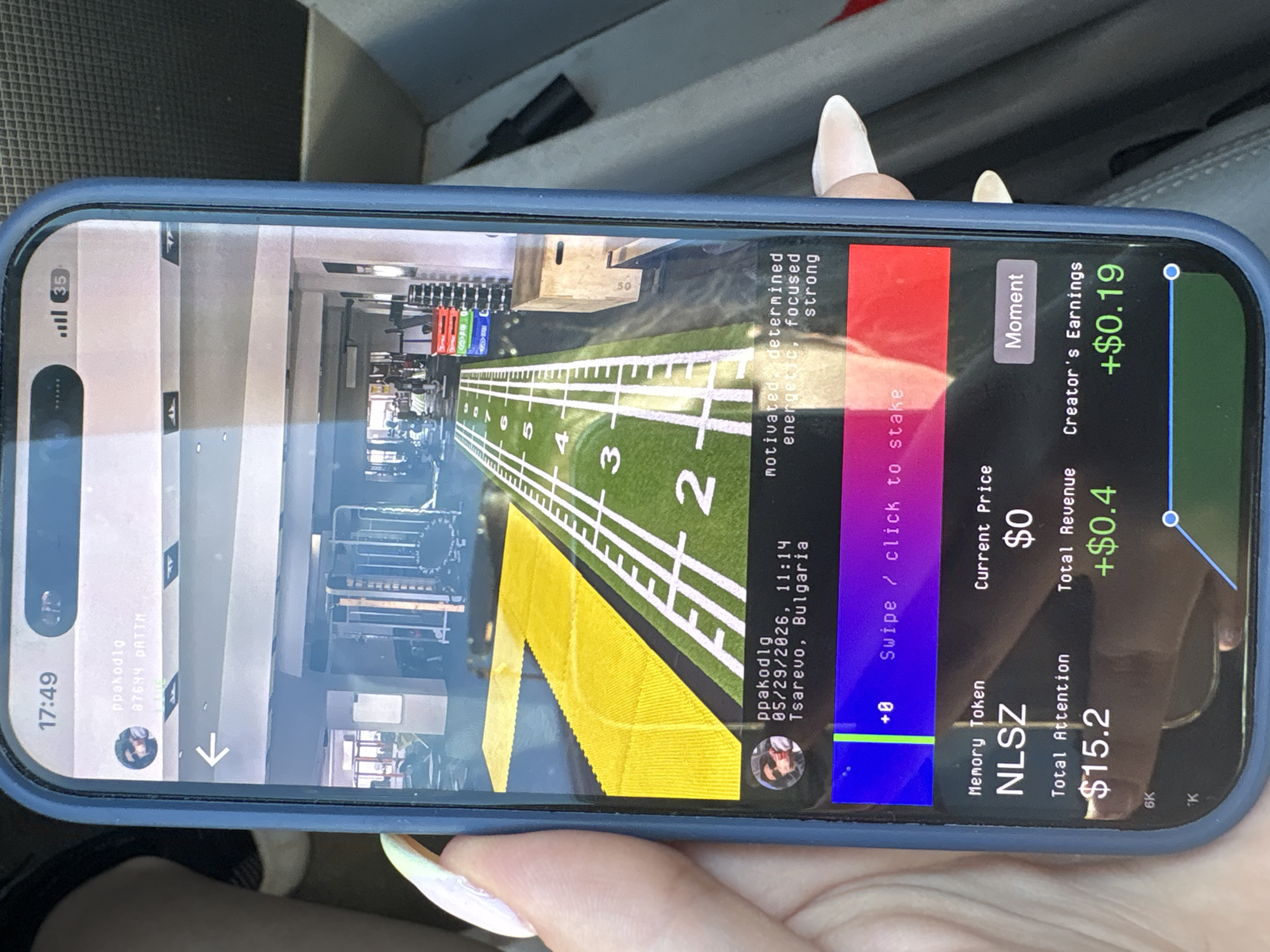 The image shows a person holding a smartphone displaying a social media app. The app features a video or image of a running track in a gym, with numbers marking the lanes. The app also displays information about a "Memory Token" called NLSZ, with a total attention value of $15.2, a current price of $0, and total revenue of +$0.4. Creator's earnings are shown as +$0.19. The user "ppakodlg" is associated with the content, and the date "05/29/2026, 11:14" and location "Tsarevo, Bulgaria" are displayed. The app interface includes buttons for "Moment" and staking. The phone has a blue case. The background suggests the photo was taken inside a car. There is no indication of the city of Burgas.