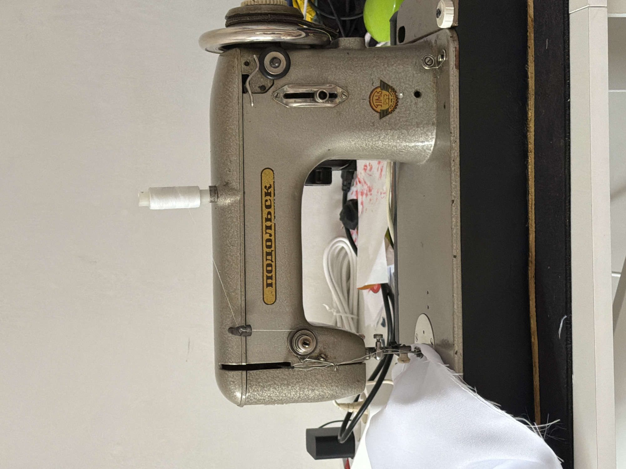 The image shows a vintage sewing machine, likely a "Podolsk" model, on a white table against a plain white wall. A spool of white thread is mounted on top of the machine. White fabric is positioned under the sewing needle, suggesting an ongoing or recent sewing operation. Various cords and other objects are visible in the background, partially obscured. The overall setting appears to be a domestic workspace. There is no indication of the city of Krasnyi Krym, Russia in the image itself.