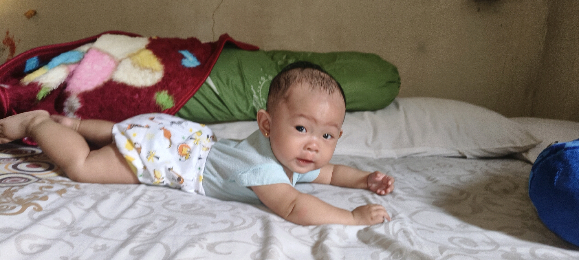 The image depicts a close-up, wide-angle shot of a baby lying on its stomach on a bed. The baby is wearing a light blue t-shirt and patterned diaper. Its head is turned towards the camera, and its eyes are open. The baby's legs are bent at the knees, and its arms are extended forward, with hands resting on the bed. Behind the baby, a green bolster pillow and a red blanket with colorful patches are visible. The bed has a white patterned sheet. The background consists of a beige-colored wall. This scene is likely captured in a domestic setting within the city of Waisai, Indonesia, given the context provided. The lighting appears to be ambient room light.