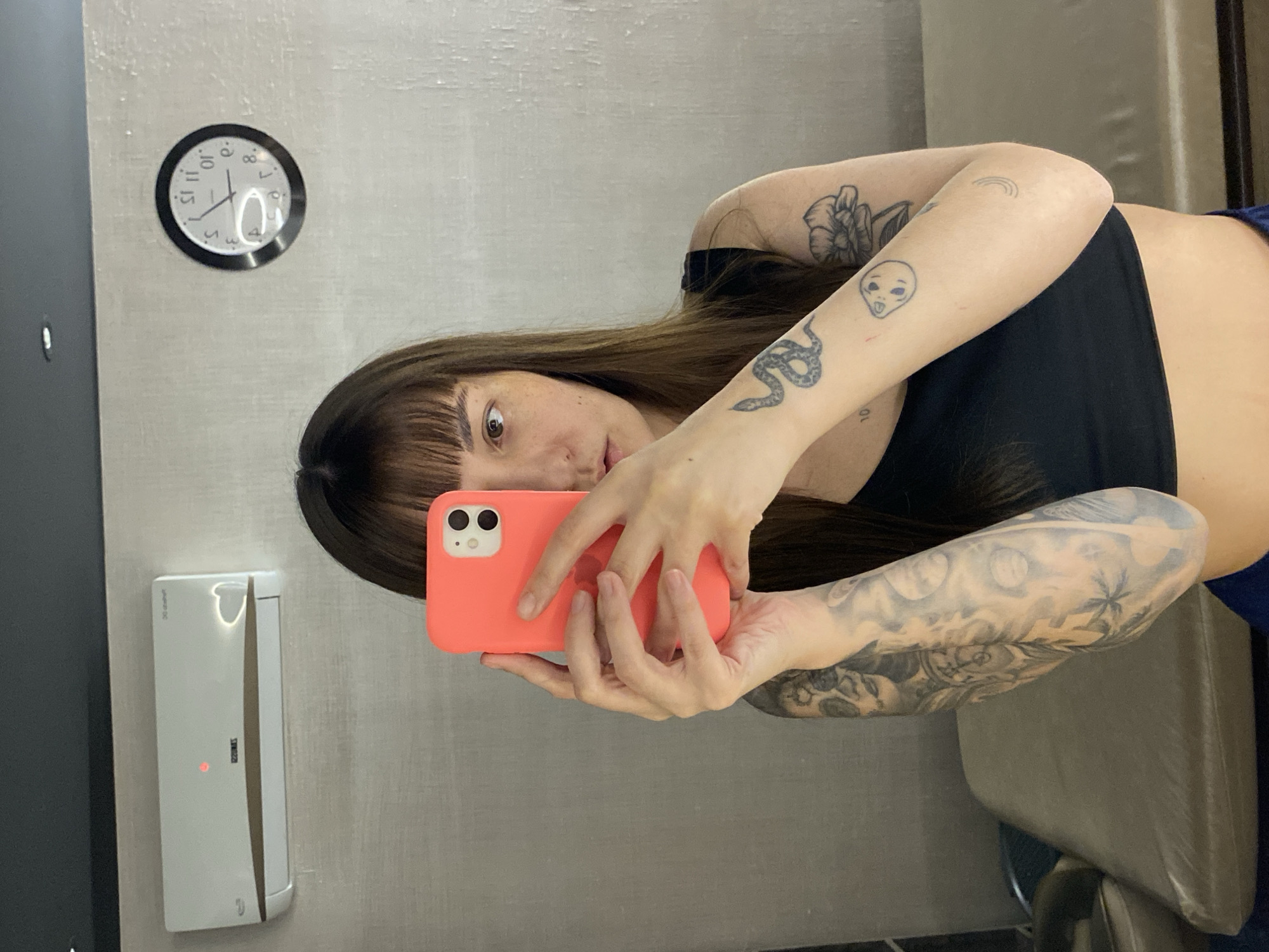 A person is captured in a mirror selfie, holding a coral-colored iPhone with both hands. The image is taken indoors, against a light-colored, textured wall. The individual has long, dark brown hair with bangs and brown eyes, looking directly into the camera with a neutral expression.
Notable details include a collection of tattoos on both arms. On the person's right arm (viewer's left), there's a detailed snake tattoo, an alien head with its tongue out, and a small rainbow. On the person's left arm (viewer's right), a larger floral design is visible near the shoulder, and a full sleeve design appears to depict celestial bodies like moons and possibly a landscape with palm trees. The person is wearing a black crop top.
In the background, a round wall clock with a black frame and white face is visible, indicating the time as approximately 9:55. The numbers on the clock face, particularly 10 and 11, appear reversed, confirming the mirror selfie. Below the clock, a white, wall-mounted unit, possibly an air conditioning unit or a smart device, features a faint red indicator light and barely legible text "ZAMESS" and "30 series". Text on the person's forearm below the alien tattoo reads "10L".
The setting is an indoor space, potentially a home or private room, in Votkinsk, Russia.