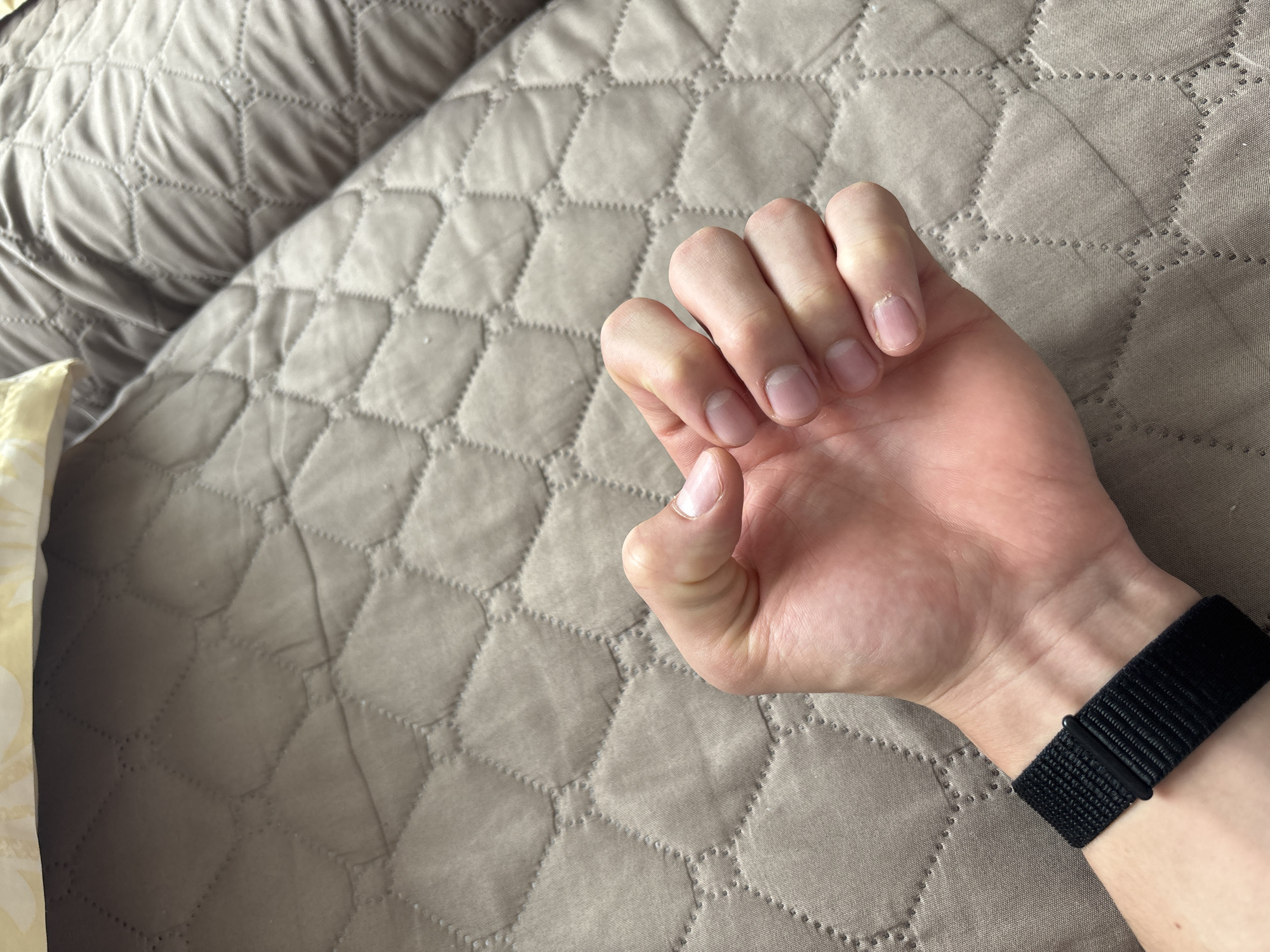 A right hand wearing a black, woven wristwatch is positioned against a quilted beige surface, potentially a bedspread or similar textile. The hand is loosely clenched, with the fingertips visible. The setting is indoors. The location cannot be determined from the image and there is no information to suggest the image was taken in Zlatograd, Bulgaria.