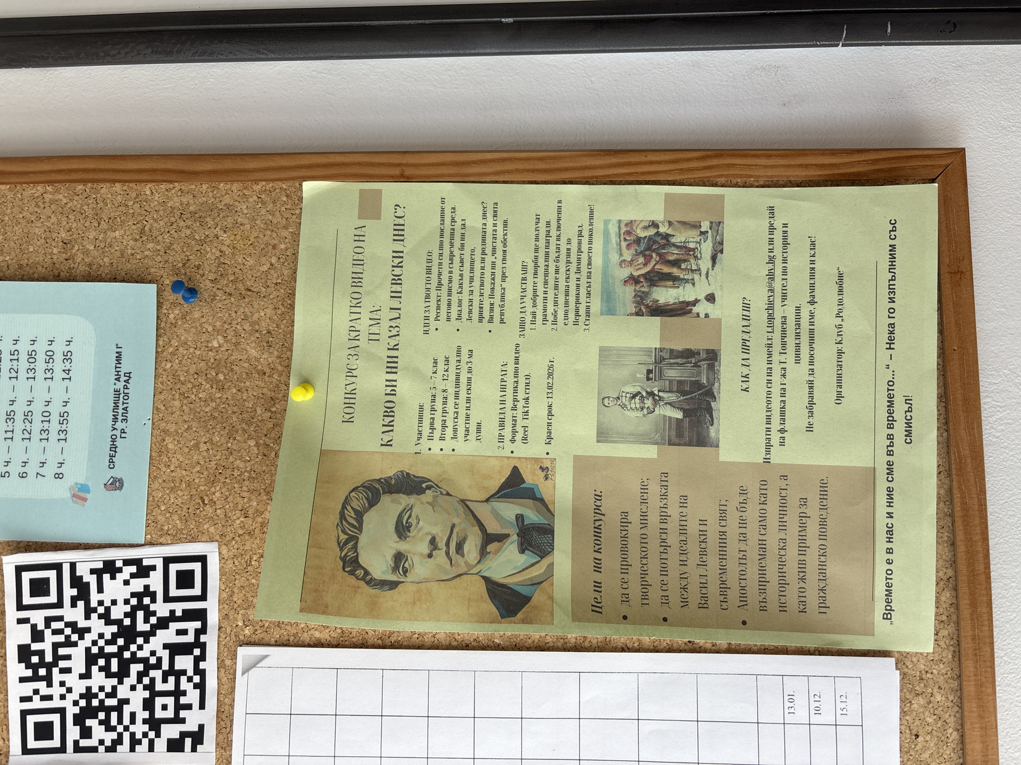 The image displays a cork bulletin board, framed in wood and mounted on a light-colored wall. Several documents are pinned to it using yellow and blue thumbtacks.
The most prominent item is a large, green-tinted flyer announcing a video competition titled "WHAT WOULD LEVSKI TELL US TODAY?". The competition focuses on Vasil Levski, a Bulgarian national hero. It is open to students in grades 5-7 and 8-12, allowing individual or team participation (up to three people). The flyer suggests video ideas such as interpreting Levski's letters in a modern context, imagining his advice on school, friendship, or homeland, or depicting his vision of a "pure and sacred republic." The required format is vertical (TikTok style), and the deadline is February 13, 2026. Prizes include diplomas, special awards, and an excursion to Perperikon and Dimitrovgrad. The competition aims to stimulate creative thinking, connect Levski's ideals with the contemporary world, and present him as a living example of civic behavior. Submissions are via email or flash drive to a history teacher. The organizer is "Club Rodolyubie," and a quote by Levski, „Времето е в нас и ние сме във времето..." - Нека го изпълним със смисъл!, is featured. The flyer also includes a stylized portrait of Levski and two small historical paintings.
To the left of the main flyer, a light blue paper displays a school timetable for the 5th, 6th, 7th, and 8th lesson periods, listing their start and end times. This paper explicitly states "СРЕДНО УЧИЛИЩЕ "АНТИМ І" ГР. ЗЛАТОГРАД" (ANTIM I Secondary School, Zlatograd), indicating the location context. Below the timetable, a black and white QR code is visible on another paper. At the bottom right of the board, a small white paper with a grid shows three dates: 13.01., 10.12., and 15.12.
The scene depicts an informational display, likely within a school in Zlatograd, Bulgaria, promoting an educational and cultural activity.