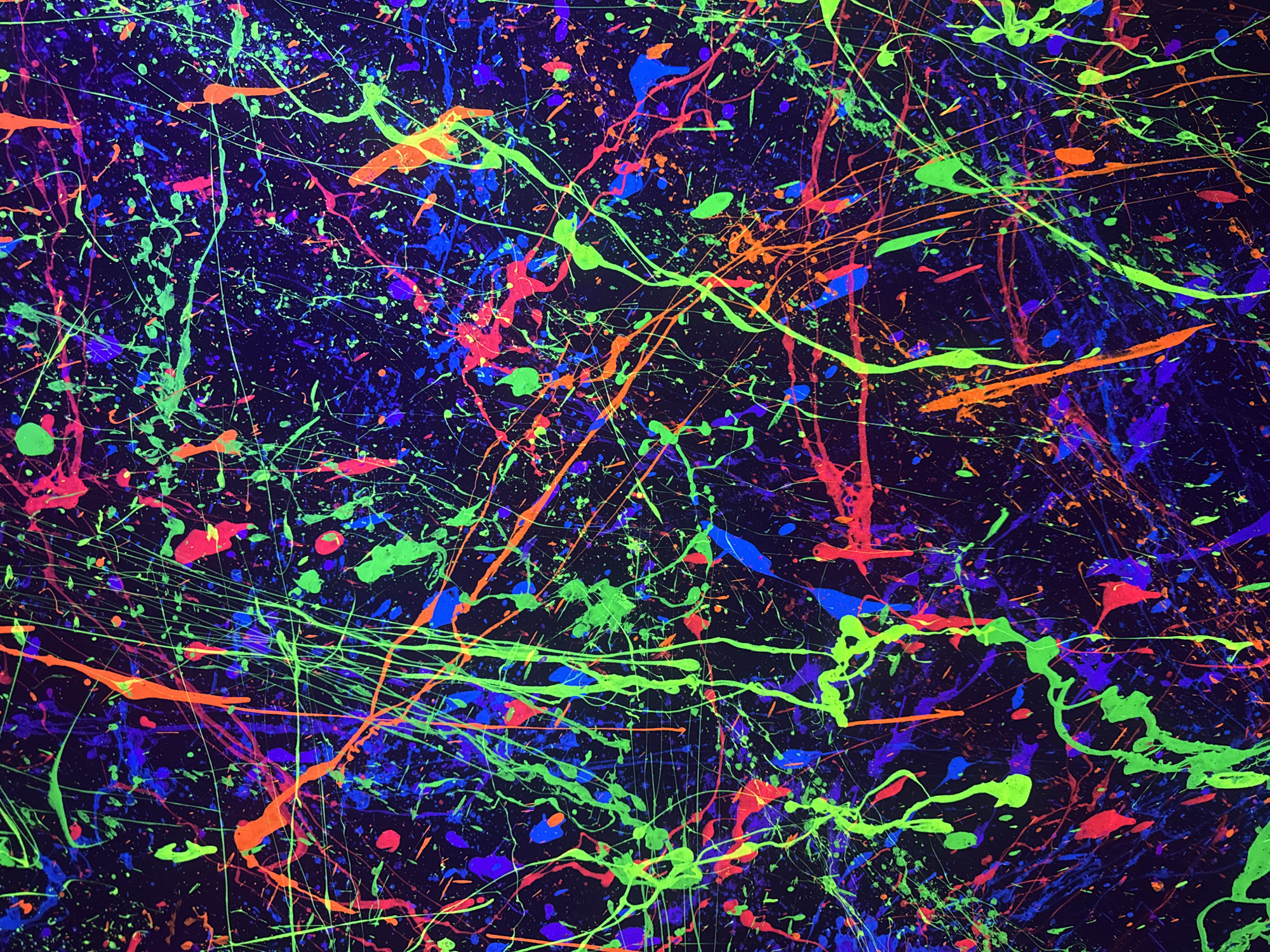 This is a photograph of a vibrantly colored abstract painting. The background is a deep, dark blue, almost black, with a textured appearance suggesting a canvas. Splattered and dripped across this background are streaks and splatters of fluorescent paint in bright neon colors, including vivid green, electric blue, hot pink, and fiery orange. These colors create a chaotic yet energetic pattern, with thin lines crisscrossing thicker splatters. The overall impression is of a dynamic, high-energy abstract composition. There is no indication of a specific location in Munich, Germany, nor are there any people, subjects, objects, actions, interactions, or environmental details that could be associated with that city. The image solely depicts the abstract artwork.