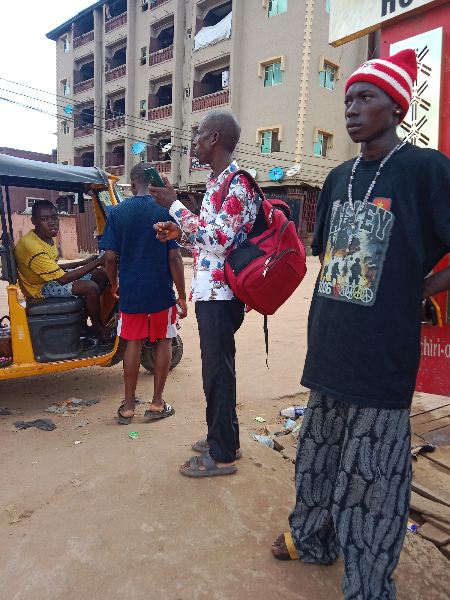 The image depicts a street scene in Nkpor, Nigeria. In the foreground, a slender man wearing a red and white striped knit cap, a black t-shirt with a graphic design, a beaded necklace, and patterned gray pants stands with his hands on his hips. To his left, a man in a floral shirt and black pants stands, holding a smartphone. Behind him is another man wearing a blue shirt and red shorts. A yellow auto rickshaw is parked on the left side of the image, with a man sitting in the driver's seat. Litter is scattered on the ground. In the background, a multi-story building is visible.
