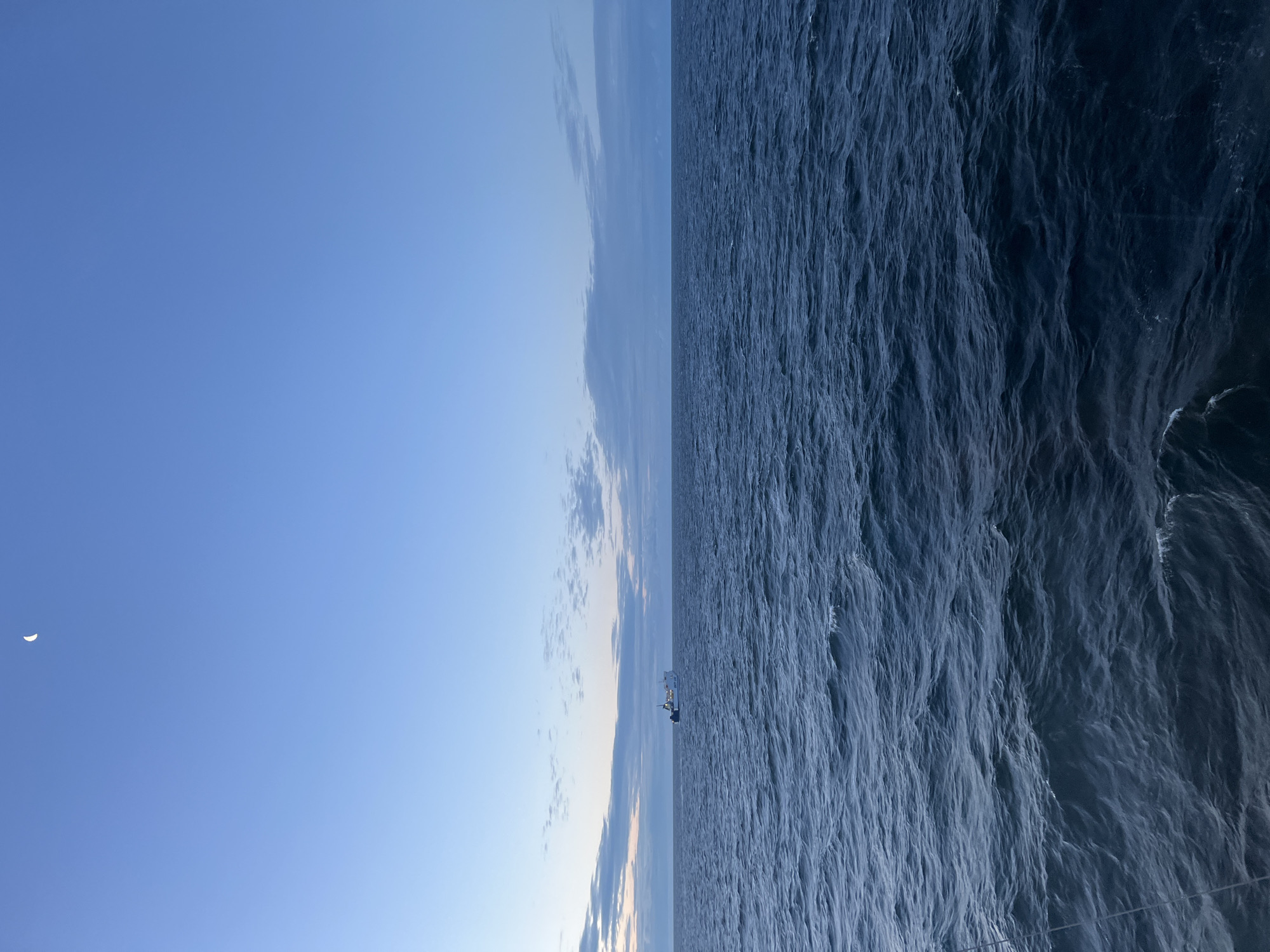 The image shows a serene ocean scene at dusk. The sky is a clear gradient of light blue at the horizon, transitioning to a deeper blue overhead, with a sliver of the crescent moon visible in the upper left quadrant. A thin band of clouds is present near the horizon. In the middle distance, a white ferry with blue accents is sailing across the dark, choppy water. The foreground is dominated by the deep blue ocean, with visible waves and ripples. The location is not specified within Poland, and there are no people or discernible human activity beyond the presence of the ferry.