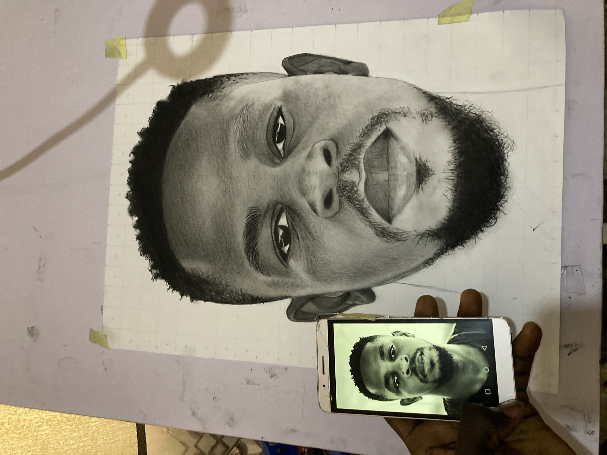 This media file shows a close-up, overhead view of a charcoal portrait of a man in progress. The drawing is on a large sheet of paper, marked with a grid of squares, suggesting a technique for accurate reproduction. The portrait is oriented upside down in the frame, with the man's head appearing near the bottom of the image and his feet at the top. The man has short, curly hair and a full beard and mustache. His eyes are open and looking directly at the viewer, and his mouth is slightly open, revealing his teeth. The shading and detail in the drawing are highly realistic, capturing the texture of the skin, hair, and the light reflecting in his eyes.
In the foreground, a hand holds a smartphone, also displaying a portrait of the same man. The phone's screen is illuminated, showing the man's face in a similar pose to the drawing, suggesting the drawing is being created from a reference photo on the phone. The hand holding the phone appears to be dark-skinned.
The setting appears to be an indoor workspace. The background consists of a light-colored wall with some faint smudges of charcoal dust. A thin yellow piece of tape is visible in the upper left corner of the drawing paper, and another piece is near the top center, holding the paper to an unseen surface. The overall lighting is somewhat dim, with a soft shadow cast across the top of the drawing paper from an overhead light source. The image evokes a sense of focused artistic creation.