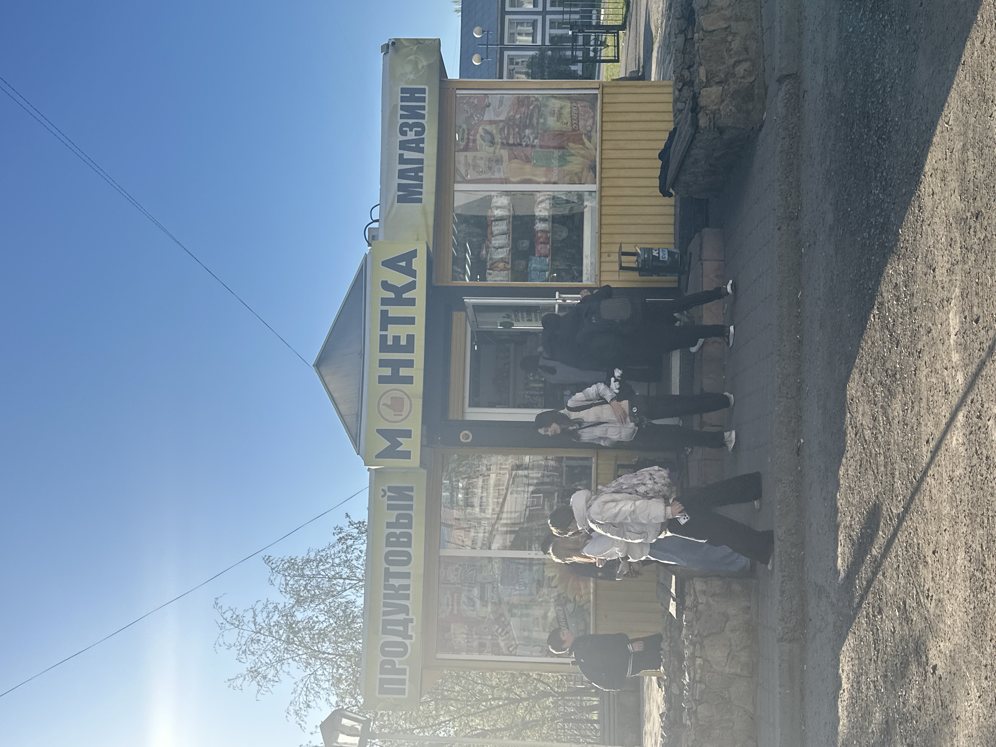 A bright, vertical, medium shot shows people waiting in line to enter a Монетка (Monetka) convenience store. The store is located in Shelekhov, Russia. The store is yellow with a black roof. The store sign reads "Monetka" and "Produktovy" in Russian. The window displays images of snacks and other goods. Six people stand in line, most wearing face masks. The street is grey and paved. Sunlight casts shadows on the ground. The sky is blue and clear.