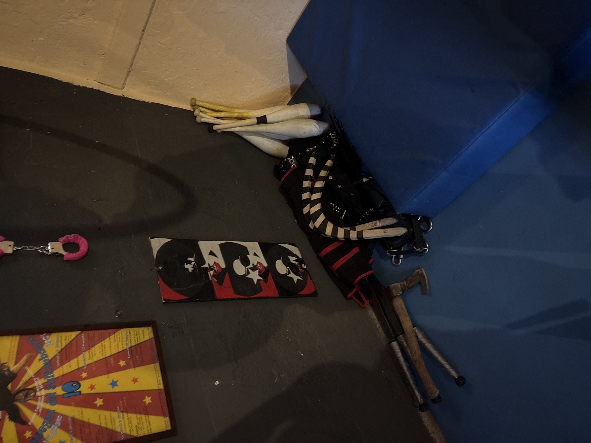 The image displays an indoor scene, likely a storage area or backroom within a performance venue in Milan, Italy. Various props and equipment are arranged on a dark gray floor, bordering a light-colored wall and a large blue cushioned mat or block.
In the upper right section, several white juggling clubs, some with yellow tips or handles, are casually stacked against the blue mat. Adjacent to these, a black item, possibly a costume piece, bag, or harness, is visible, featuring silver buckles and black-and-white striped elements. Below this, three varying walking sticks or crutches, made of wood and metal, are laid out on the floor. One has a distinctive curved handle, reminiscent of a vintage walking stick.
To the left, a pair of pink, toy-like handcuffs connected by a silver chain rests on the floor. Below and slightly to the right of the handcuffs, a flat, rectangular object, possibly a banner or box lid, showcases a repeating graphic pattern. This pattern features stylized black and white cartoon-like skull or jester faces with red accents, set against a black background.
Further down and to the left, a framed piece of artwork or a poster lies on the floor. It features a vibrant red and yellow starburst design adorned with multiple small stars. The number "10" is prominently displayed in the center of this poster. While other text is present on the poster, it is largely unreadable due to blur and perspective.
The scene depicts equipment commonly associated with circus arts, magic shows, or theatrical performances, suggesting a setting either before or after an event. There is no direct activity taking place, and the lighting is typical of an indoor environment, making the time of day indeterminate. No specific emotions are conveyed.