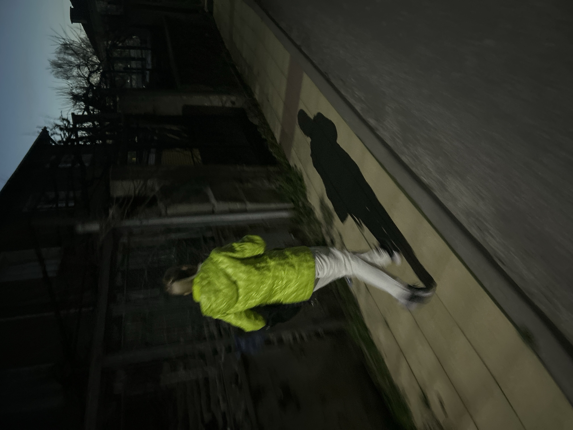 A person wearing a bright green jacket and white pants walks on a sidewalk next to a building. The photo appears to have been taken at night. The building is dark and partially obscured by trees or overgrown vegetation. A shadow is cast on the wall adjacent to the sidewalk. The location is possibly Zlatograd, Bulgaria, as specified in the prompt.