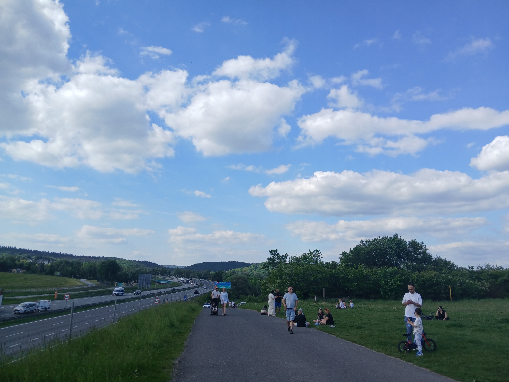 The image shows a group of people gathered on a paved path and grassy area adjacent to a highway. The location is in Unknown, Unknown. The highway is to the left, with multiple lanes of traffic visible. There are vehicles of various types moving along the highway. A line of trees and bushes separates the path and grassy area from the highway. People are walking along the path, sitting in the grass, or standing around talking. Some are using mobile phones. The sky is blue with numerous white clouds. In the background, there are rolling hills covered with trees. The overall scene suggests a recreational or viewing area near a highway.