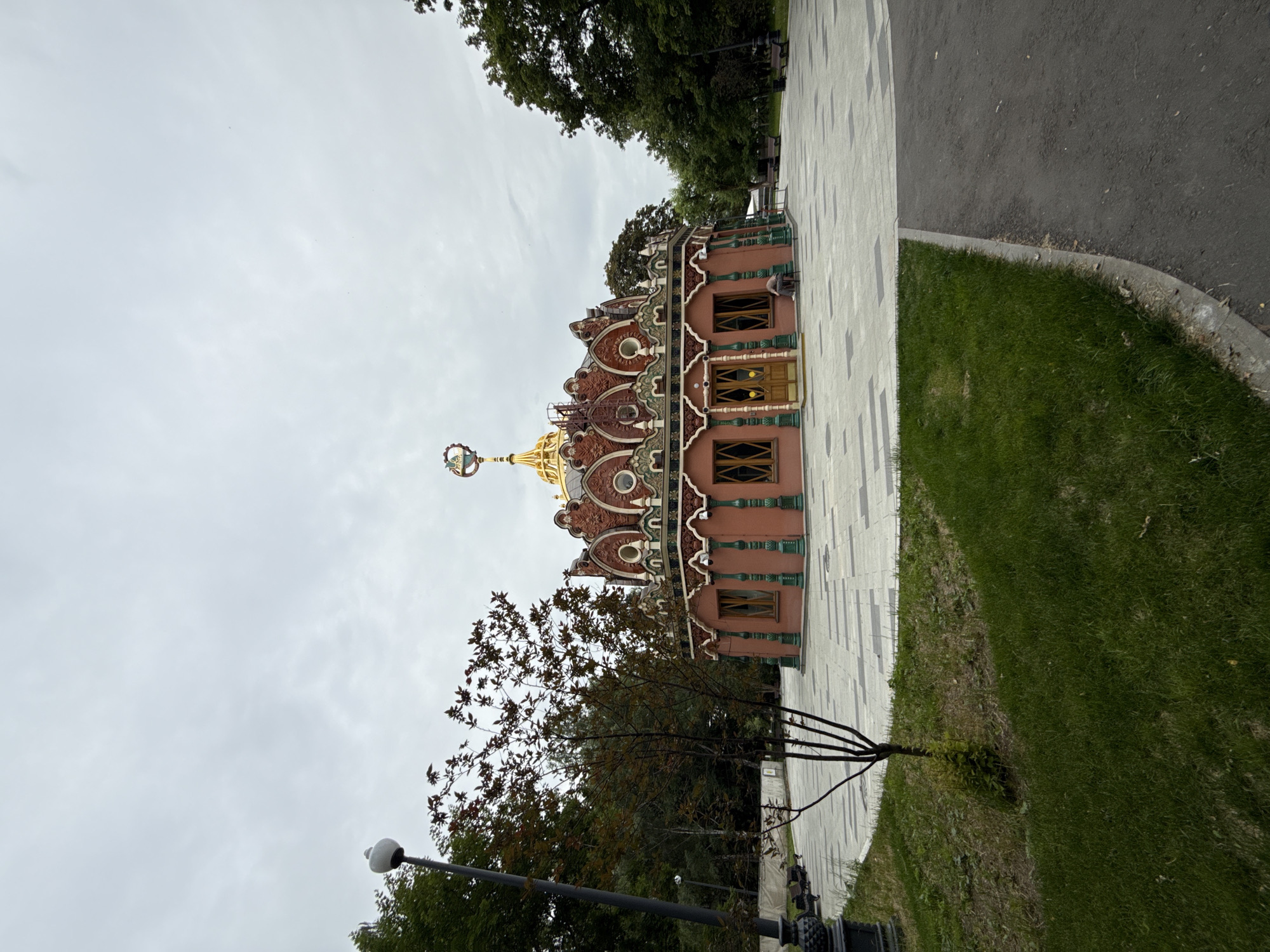 The image depicts a building with ornate, traditional Russian architecture, featuring a gilded dome and intricate red and green decorative elements. The building appears to be a small pavilion or kiosk, possibly a historical monument or a unique public structure. It's situated on a paved walkway, with a patch of well-maintained grass and a dark asphalt path adjacent to it. Mature trees frame the scene, some with dark green foliage and others appearing to be deciduous with hints of autumnal colors. The sky is overcast, suggesting a cloudy day, and the lighting is diffuse. A lamppost with a white globe is visible in the foreground. The overall impression is one of a peaceful, urban park or garden setting in Moscow, Russia, possibly on a slightly cool day. No people are actively engaged in any discernible activity, though a person can be seen sitting on a bench in the background. The focus is on the architectural details of the building and the serene landscape.