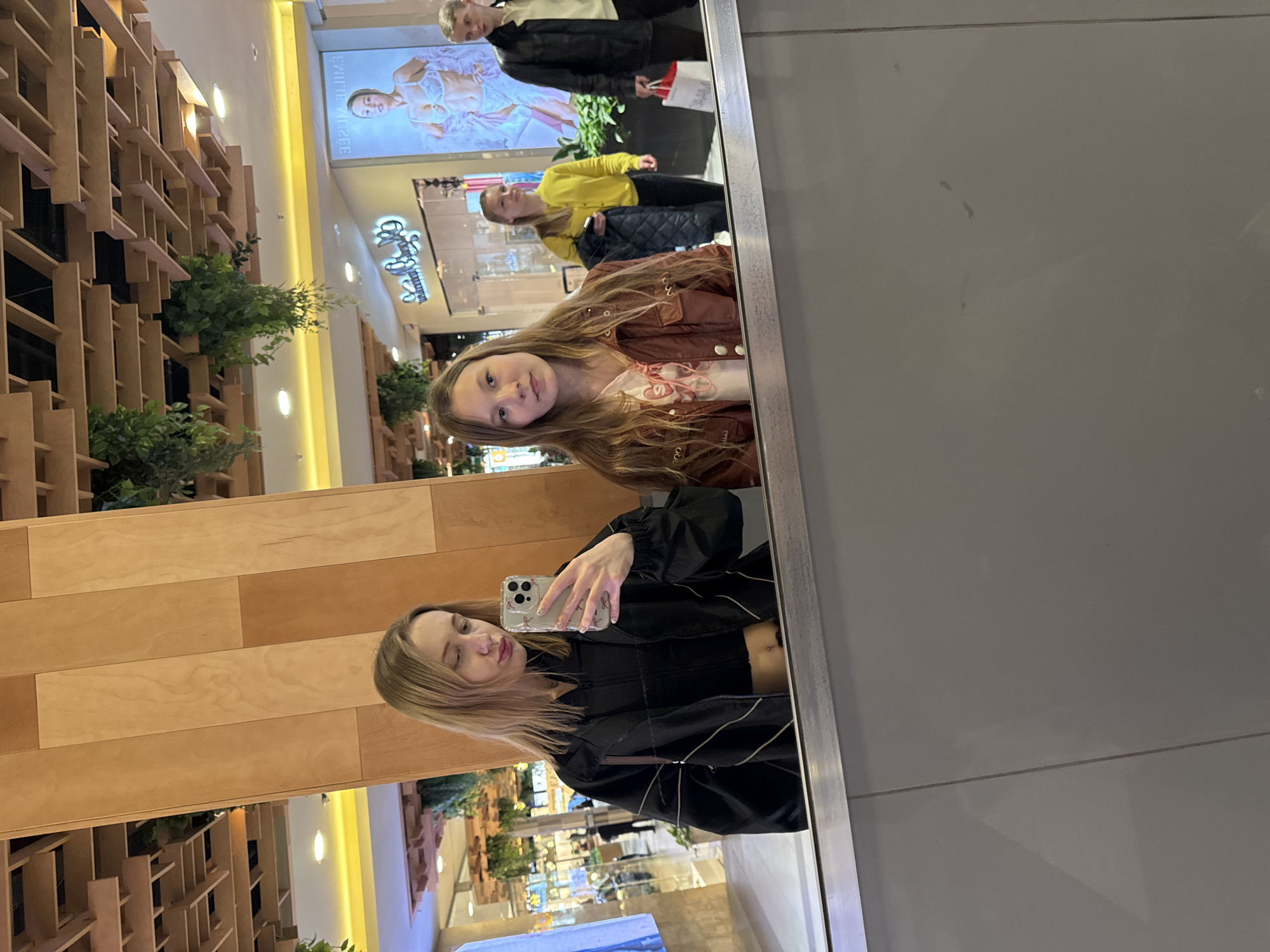 The image depicts a woman taking a selfie in a mirror located in a shopping mall, which can be identified as being in Ol'ginskaya, Russia, based on mall features in reflections. The mirror reflects herself and a younger girl behind her. The woman holds a smartphone and wears a black jacket over a cropped top. The girl behind her wears a brown jacket. Other shoppers are also visible in the background. Mall interior details, including ceiling lights, signs, decorations, and store displays, are visible in the mirror and the direct view of the scene.