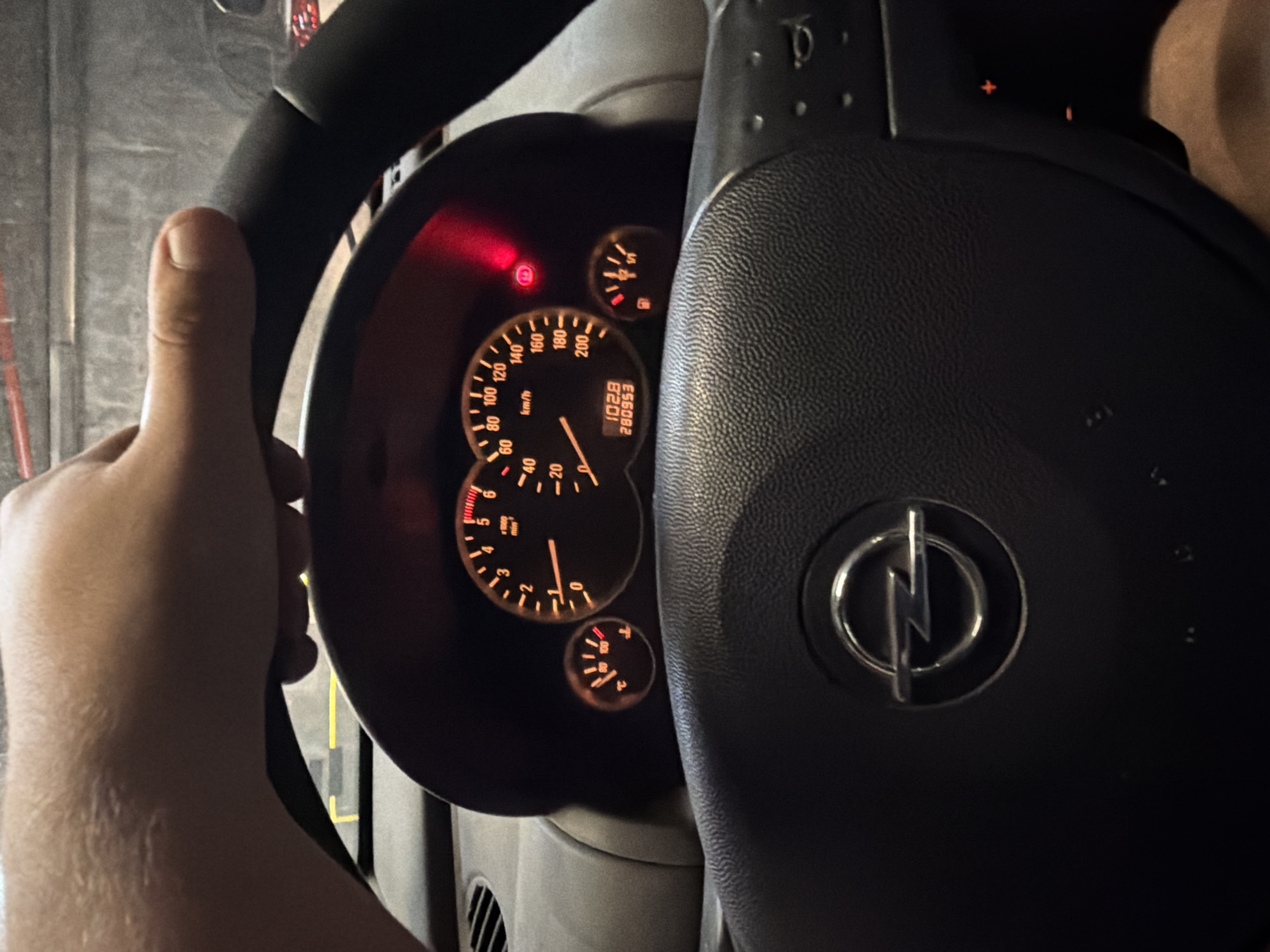 The image shows the interior of a car, specifically the steering wheel and dashboard. The steering wheel, on the right, is black with the Opel logo in the center. On the left, a hand is visible holding the steering wheel. The dashboard features a speedometer, tachometer, fuel gauge, and engine temperature gauge. The odometer displays 280953 kilometers, and the radio frequency is 102.8. A warning light is illuminated on the dashboard. The image was captured indoors, likely in a garage. It is impossible to determine the exact location within Sofia, Bulgaria, without additional context.