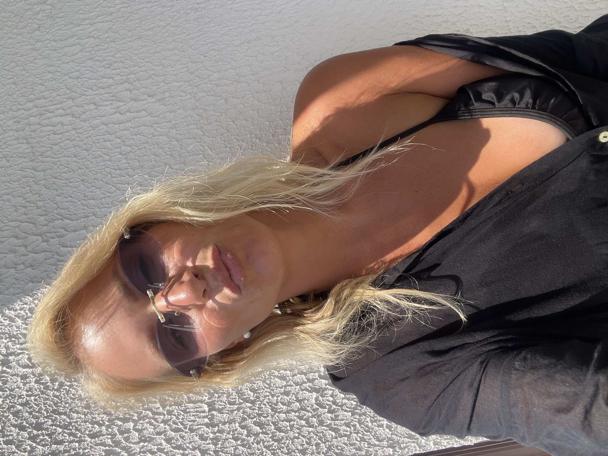 A blonde woman with fair skin, wearing sunglasses and a black top, poses for a selfie against a textured white wall. Sunlight casts shadows on her face and shoulder. She is outdoors, possibly on a balcony or patio, given the wall's texture and outdoor lighting. No specific details about Giachrypsh, Abkhazia are visible.