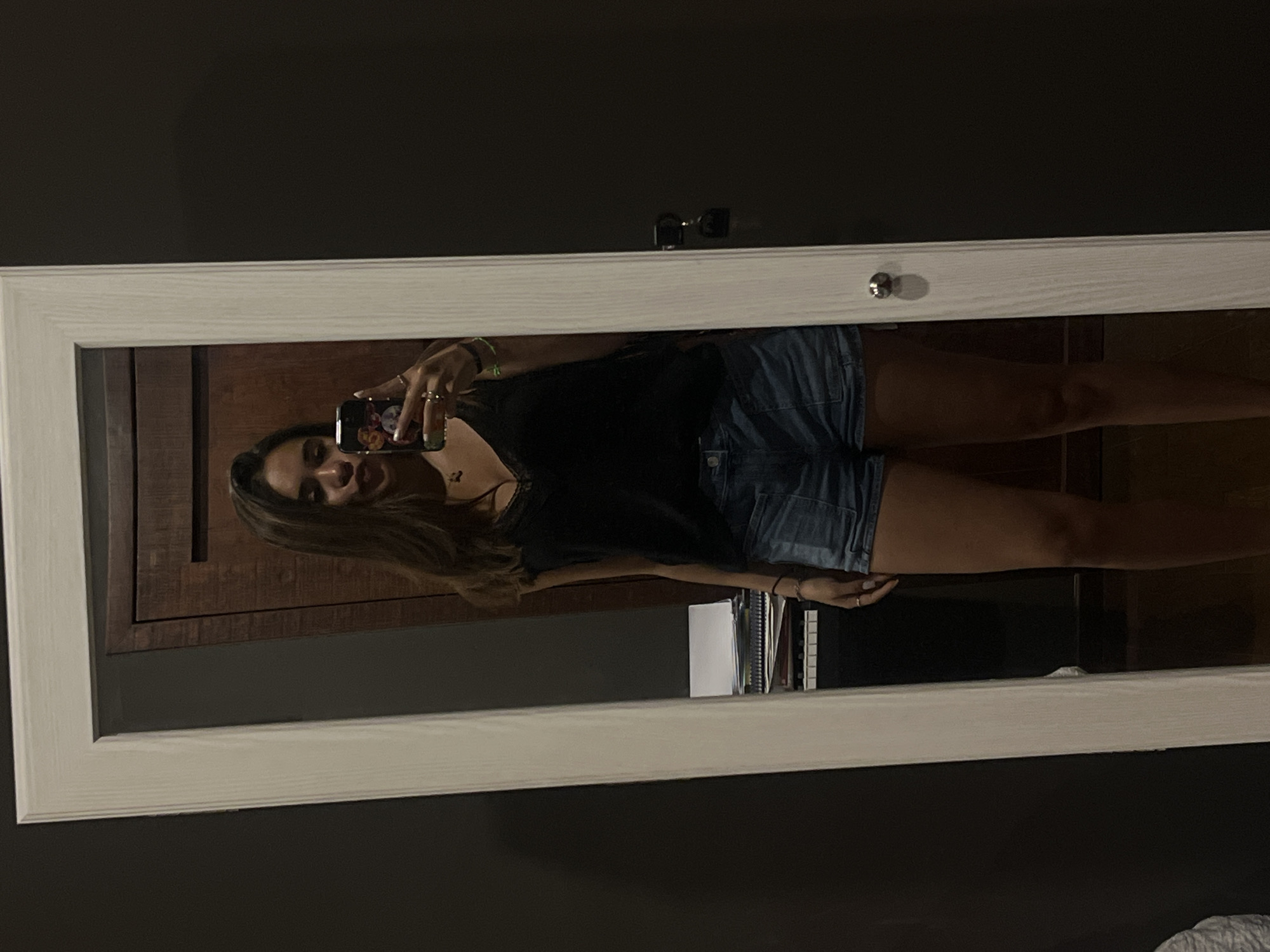 A young woman is taking a selfie in a mirror with a white frame. She is wearing a black top and denim shorts. The mirror is hanging on a dark wall and reflects a scene with a wooden cabinet and some books or notebooks. The location is not discernible from the image.