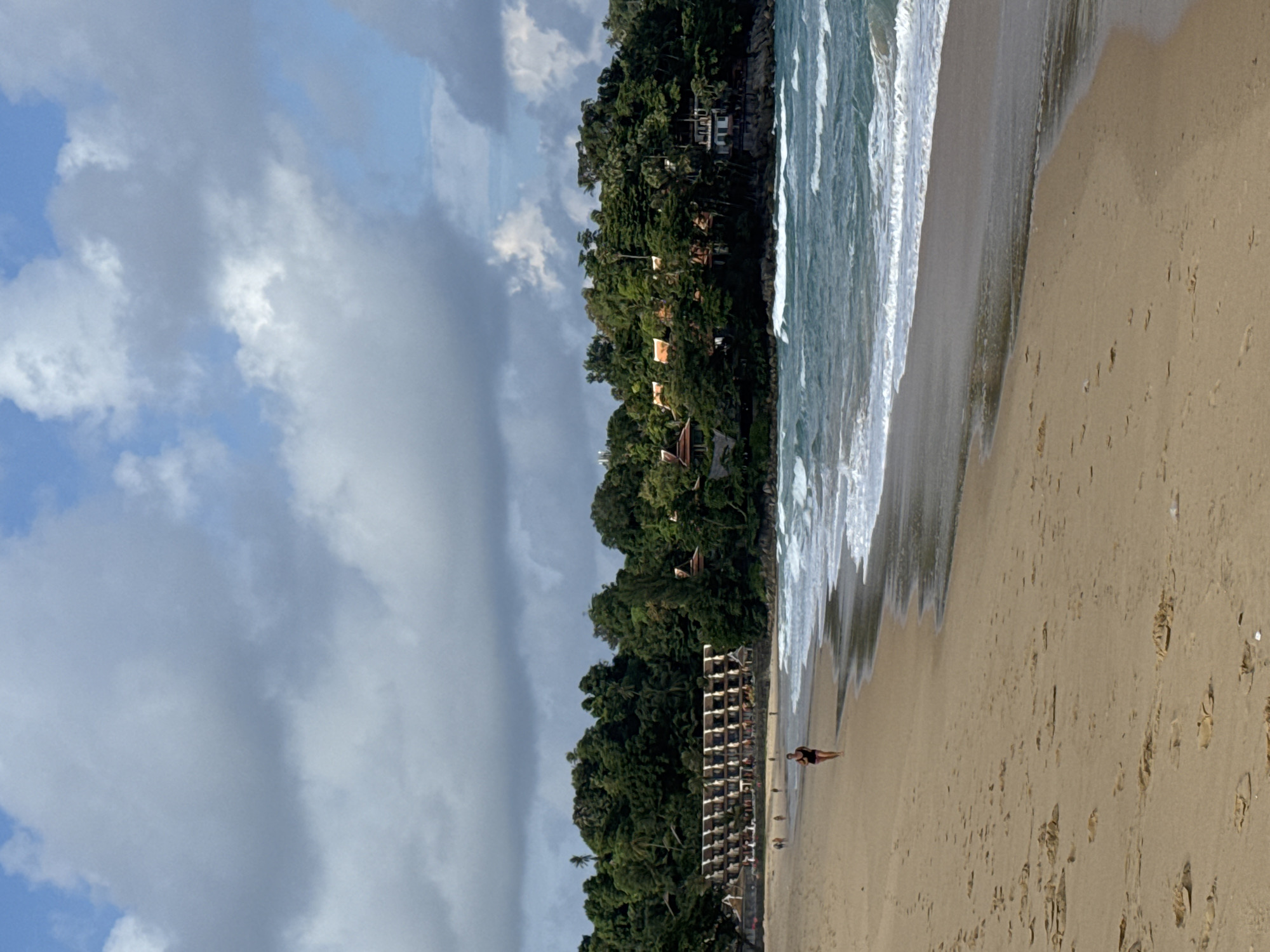 The image depicts Karon Beach in Karon, Thailand, on a cloudy day. The beach is sandy with some footprints visible. A lone woman in a black swimsuit is jogging on the wet sand near the water's edge. The ocean has moderate waves with whitecaps. In the background, a long building with multiple balconies is nestled amongst dense, green tropical foliage. Further in the distance, more trees and buildings with terracotta roofs are visible on a hill overlooking the beach and sea. The sky is a mix of blue patches and large, grey clouds.