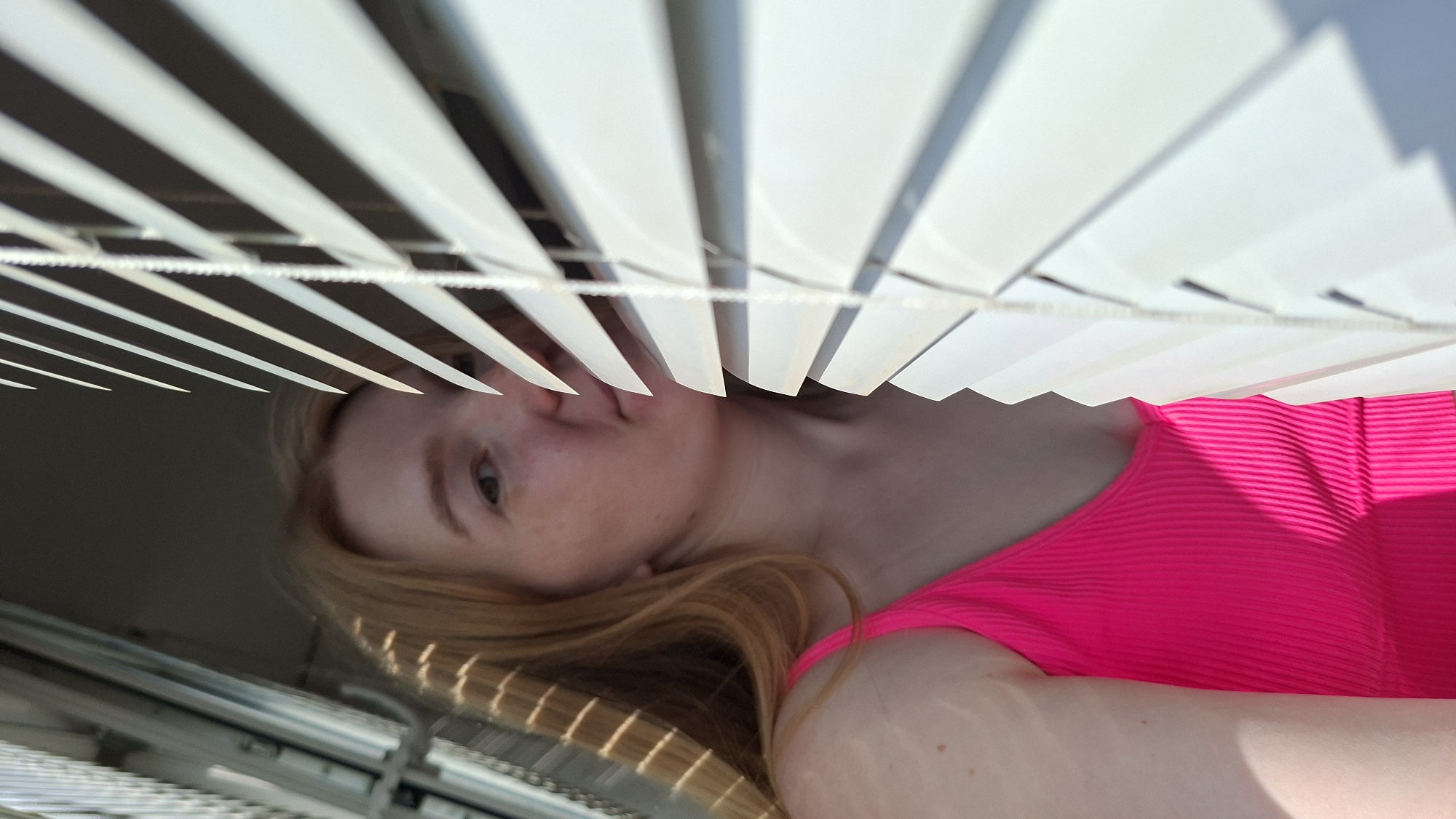 A medium shot captures a person with fair skin, freckles, and reddish-blonde hair looking down from behind white window blinds. The person is wearing a bright pink ribbed top. The background is blurred. The image is oriented in a way that suggests it was taken from below. No specific location in the city of Chaikovsky, Russia can be determined from the image.