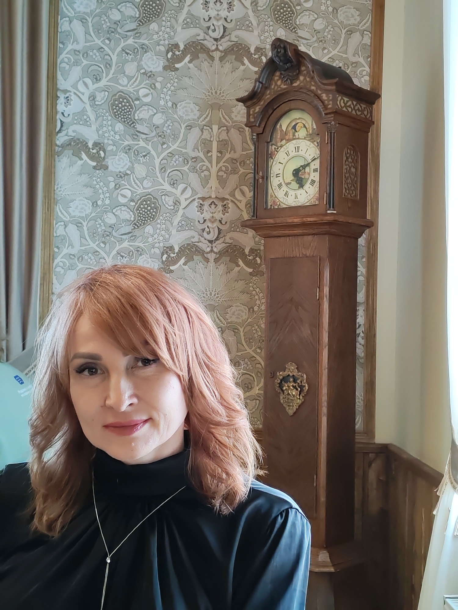 A portrait shot of a woman in front of a grandfather clock and patterned wallpaper. The woman has reddish-blonde hair and is wearing a black turtleneck and a silver necklace. The grandfather clock is wooden with a decorative top and a gold emblem on the base. The wallpaper has a floral and animal pattern. The scene is located in the city of Sarapul, Russia.