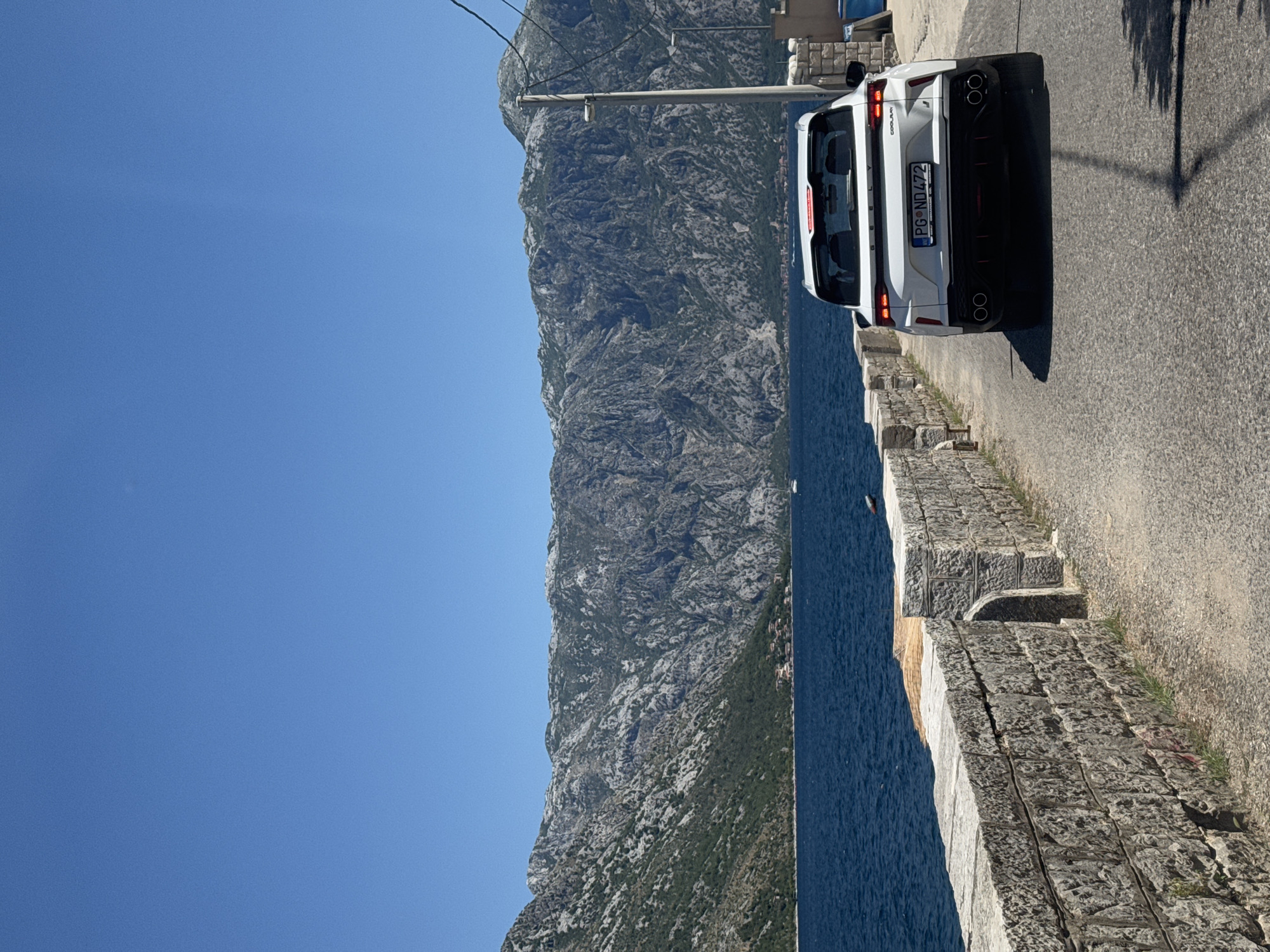 A white Geely Coolray SUV with license plate "PG ND472" drives on a paved road in Prčanj, Montenegro. The road is bordered by a weathered stone wall on the left, which runs alongside a body of deep blue water. In the background, steep, rocky mountains with patches of green vegetation rise dramatically under a clear blue sky. The car is positioned in the right lane, facing away from the camera. The scene is bathed in bright sunlight, casting shadows on the road.