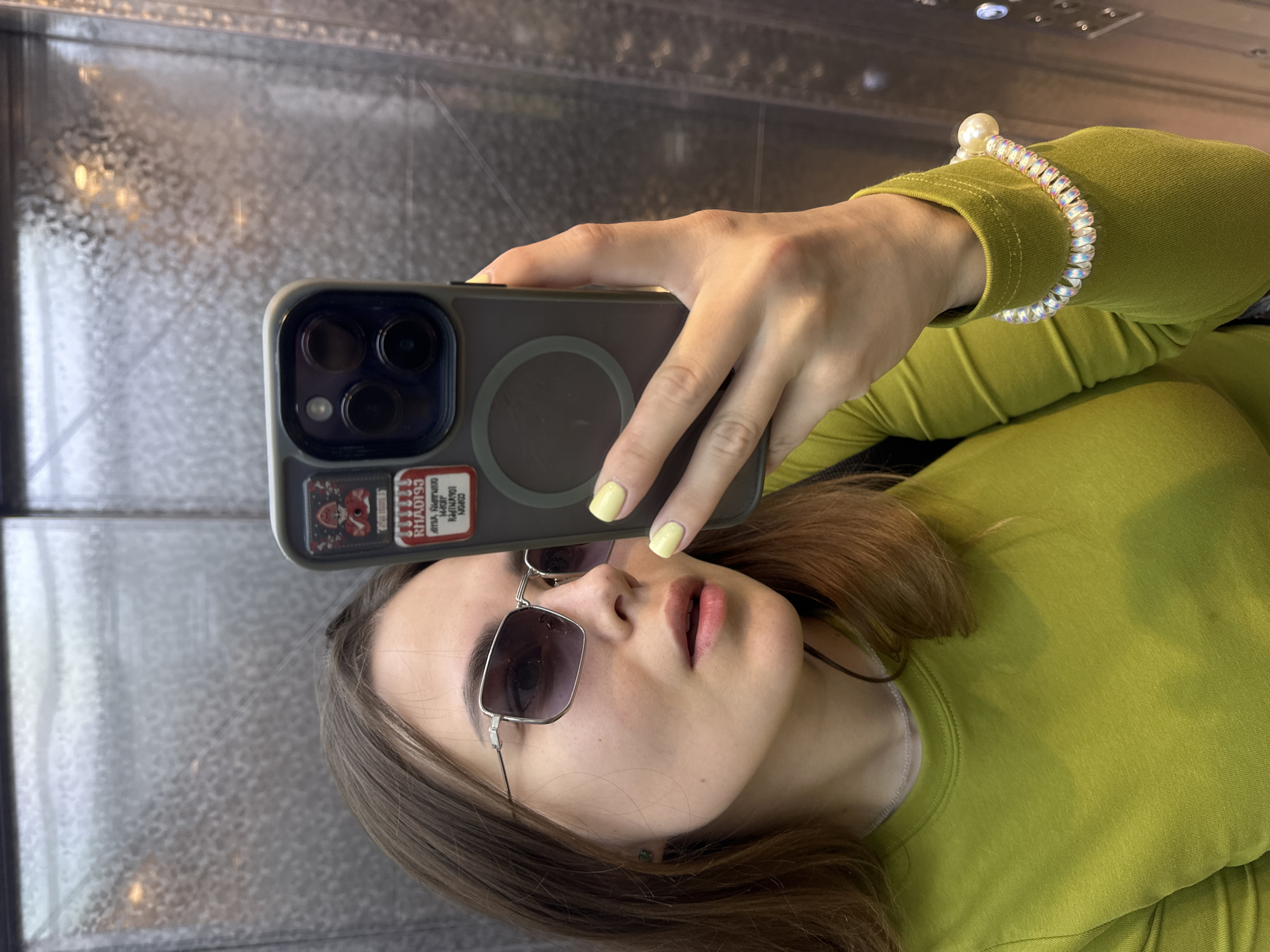 A young woman with shoulder-length brown hair and light skin is taking a selfie in an elevator. She is wearing rectangular sunglasses with purple lenses, a lime green long-sleeved shirt, and a pearl bracelet on her right wrist. She has light yellow nail polish. A sticker is affixed to the back of her phone case, which appears to be dark gray. The elevator walls are metallic and reflective, with a textured pattern. The image is taken from a slightly low angle. There are no discernible landmarks or signs indicating the specific location within the city of Perm, Russia.