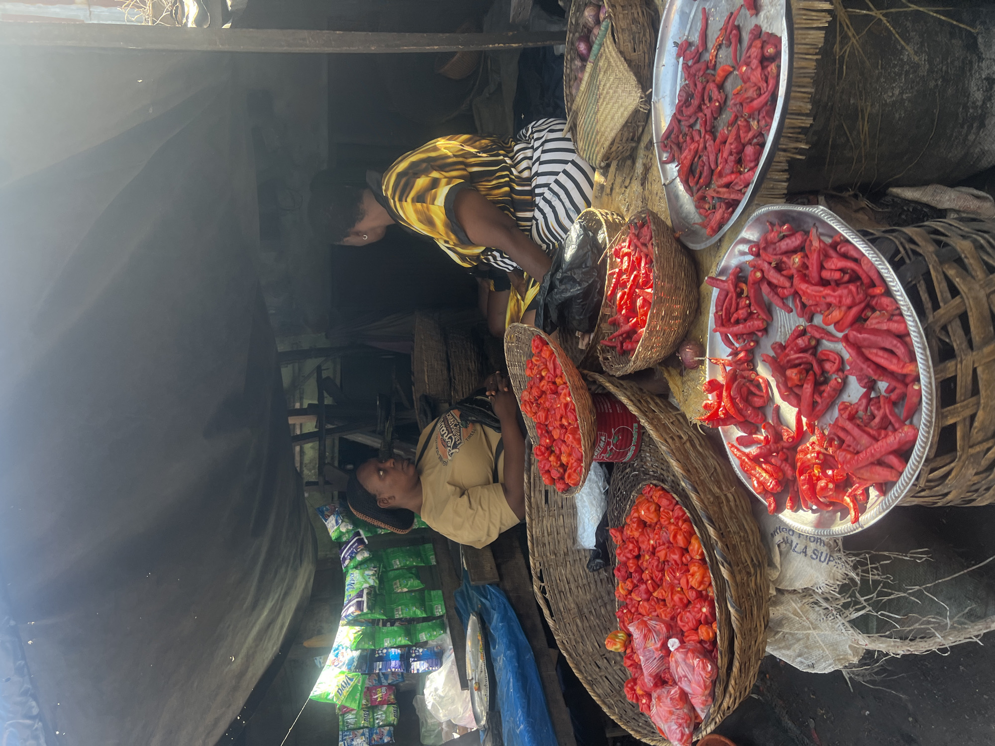 In Nkpor, Nigeria, two women are positioned at a market stall, surrounded by baskets and trays filled with red chili peppers. One woman, dressed in a yellow and black striped top, appears to be sorting or selecting the peppers. The other woman, wearing a tan-colored shirt, is partially obscured and positioned near shelves stocked with packaged goods. The stall has a makeshift covering and is open to the surrounding market environment.