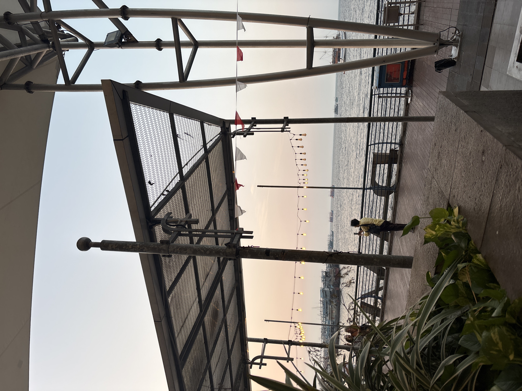 The image depicts an outdoor waterfront scene, likely in North Jakarta, Indonesia, at dusk. A man in a yellow shirt stands on a wooden boardwalk, holding a phone to take a photo of the sea. In the background, several boats are anchored. The boardwalk is covered by a metal-framed canopy with mesh infill, and decorative string lights are suspended overhead. The sea is calm, and the sky displays a gradient of light orange and pale blue, indicative of sunset. Lush green plants are visible in the foreground. Several poles and structural metal supports for the canopy are prominent features of the scene.