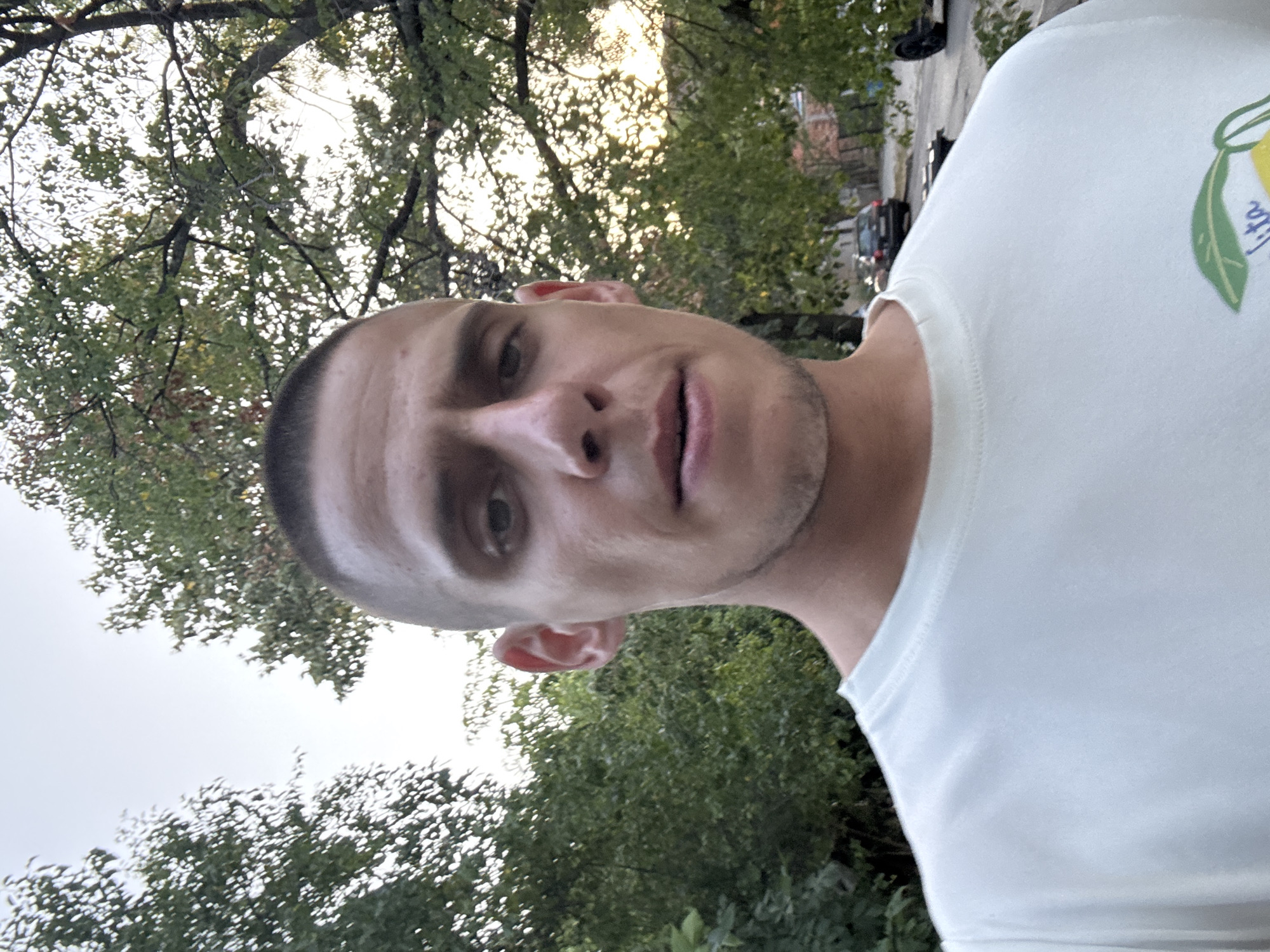 A man with a buzz cut is taking a selfie outdoors. He is wearing a light blue t-shirt with a graphic of lemons and leaves on the lower right side of the shirt. The background consists of green trees and a cloudy sky. In the distance, a street with parked cars and buildings is visible. The lighting suggests it is either early morning or late afternoon. The location is likely a residential area in Sofia, Bulgaria, given the trees and architecture visible.