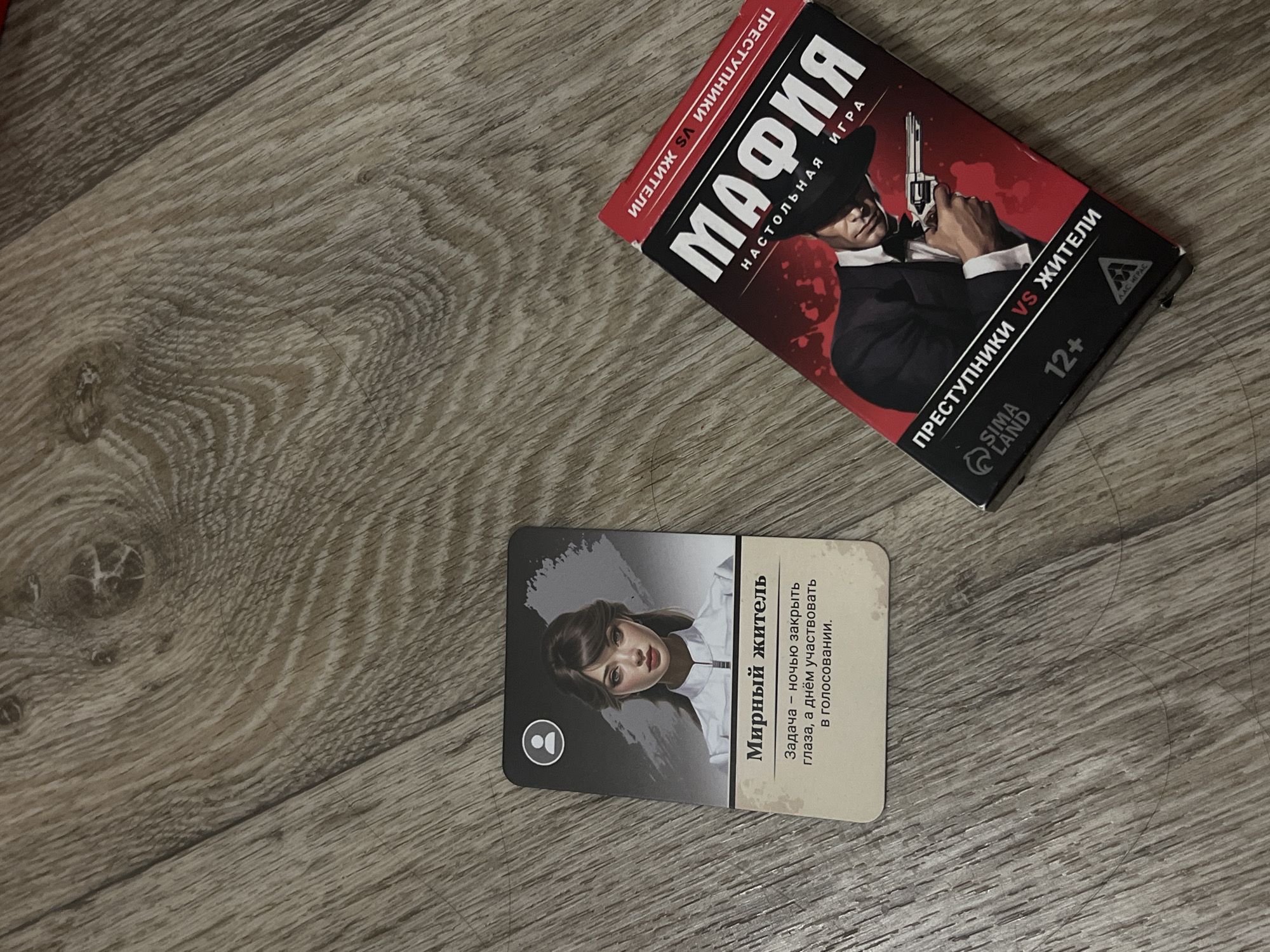 In a close-up shot, two game items are laid out on a textured wooden surface, resembling a floor. On the left, a rectangular card displays an illustration of a young woman with dark hair and a white collared shirt, her eyes closed. Below her image, the Russian text "Мирный житель" (Peaceful resident) is printed, followed by a description of her role in the game. To the right of the card, a red and black box, likely for a board game, is partially visible. The box features a dramatic illustration of a man in a suit and fedora holding a pistol, with the Russian title "МАФИЯ" (Mafia) prominently displayed in large white letters. Other Russian text on the box indicates it is a board game. The wooden surface exhibits a wood grain pattern with visible lines and knots. The overall scene is indoor, with no indication of a specific location within the city of Дачное, Russia.