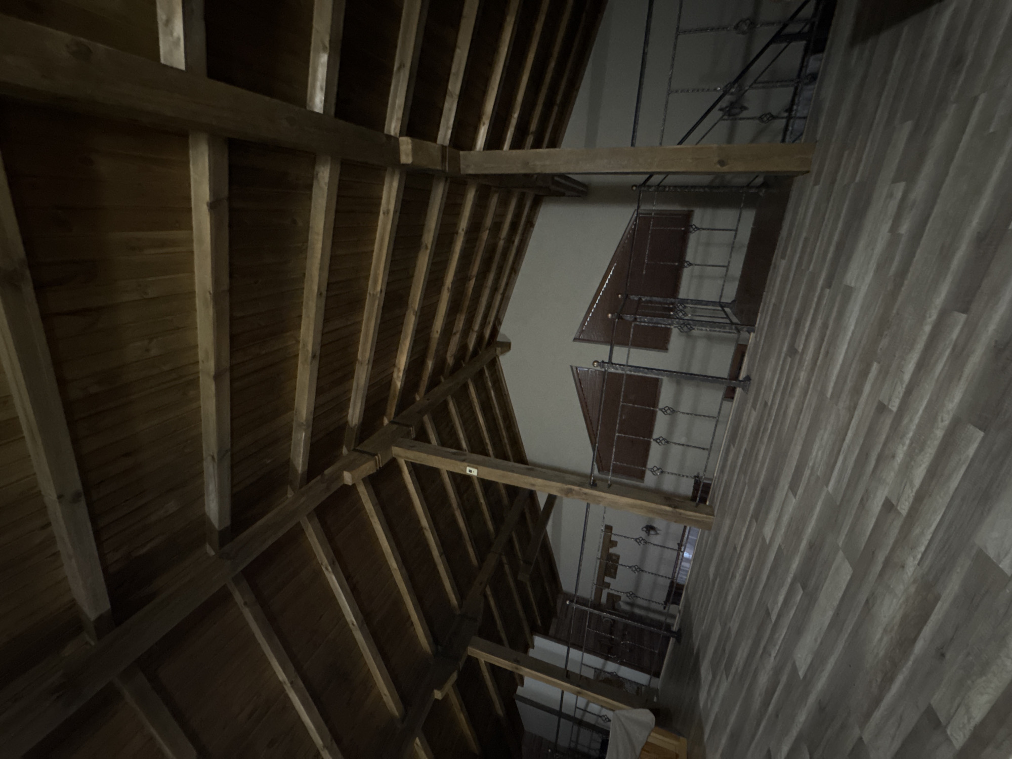 The image shows an interior view of a building, focusing on the transition between a wooden-beamed roof and a stairwell. The roof construction consists of parallel wooden planks supported by beams. The adjacent wall is covered in light-colored, wood-grained paneling. A metal railing extends along the stairwell, with visible stairs and landings. There are no people visible. The location is Dolni Marian, Bulgaria.
