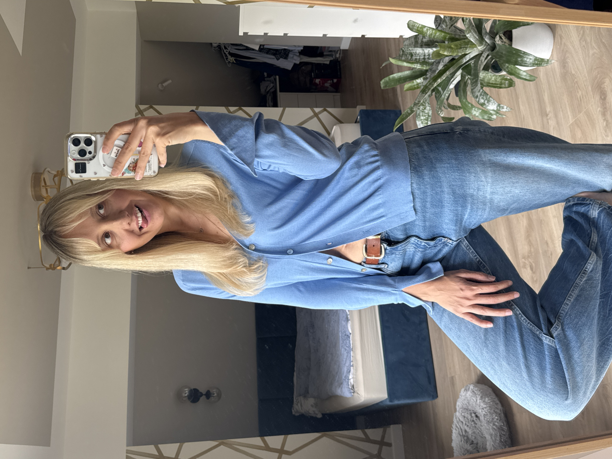 A blonde woman with bangs is taking a mirror selfie in what appears to be a bedroom. She is wearing a light blue cardigan unbuttoned to reveal her midriff and blue denim jeans. She is sticking her tongue out and looking to the side. In the background, a bed with blue bedding is visible, along with a closet full of clothes and a potted plant. The decor includes a gold geometric pattern on the wall. The overall lighting suggests it is daytime. The image does not provide any information or visual cues that would place the scene in Carnikava, Latvia, or any specific city.