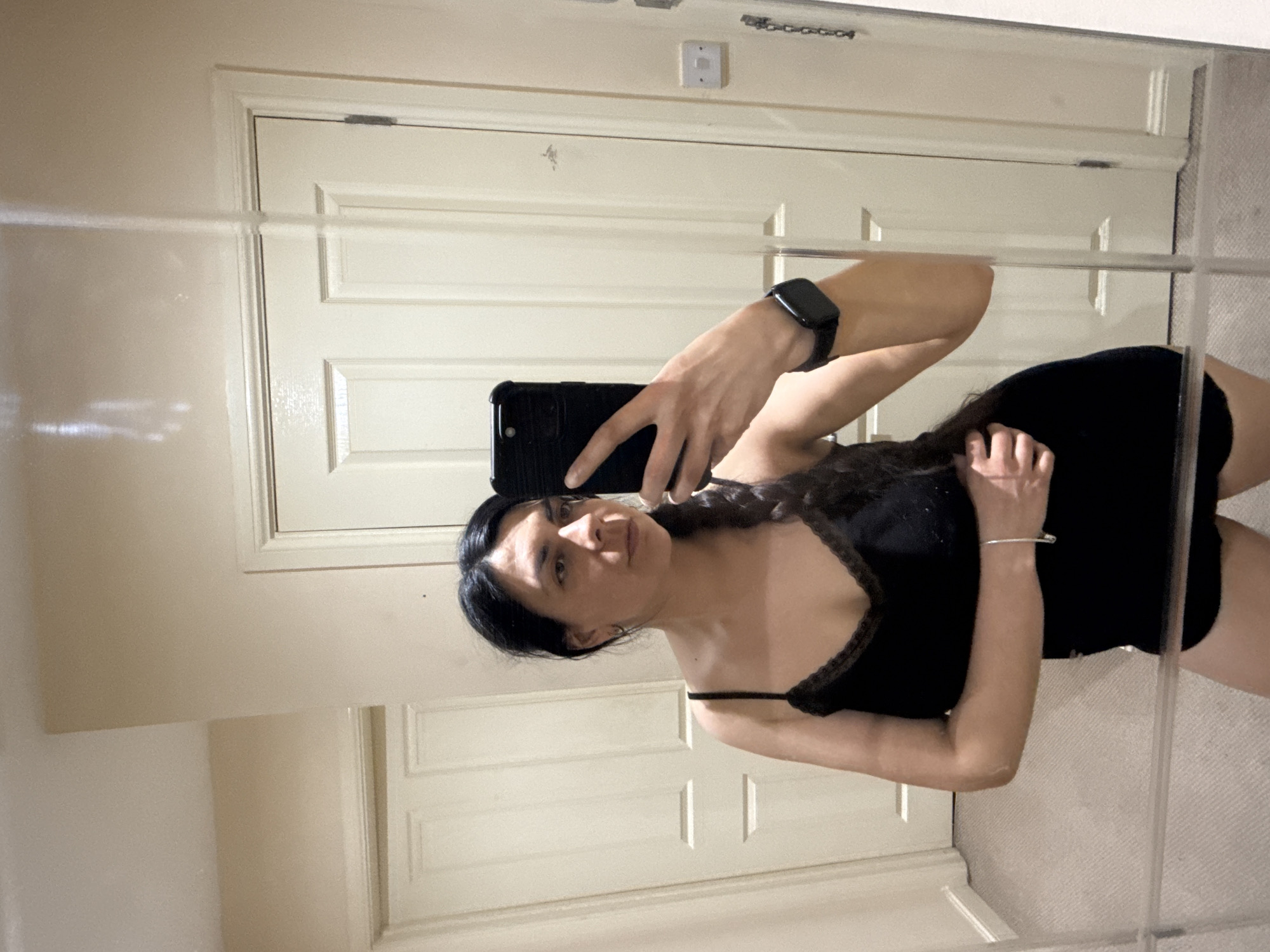 A woman stands in front of a mirror, holding a black smartphone up to take a selfie. She is wearing a black spaghetti strap top and a dark bottom garment, likely a dress or shorts. A watch is visible on her left wrist. Her dark hair is styled with a braid.
The background shows a white paneled door, part of a room in a house. The location is not discernible from the image. There is nothing visible to indicate that the image was taken in Great Yarmouth, United Kingdom.
