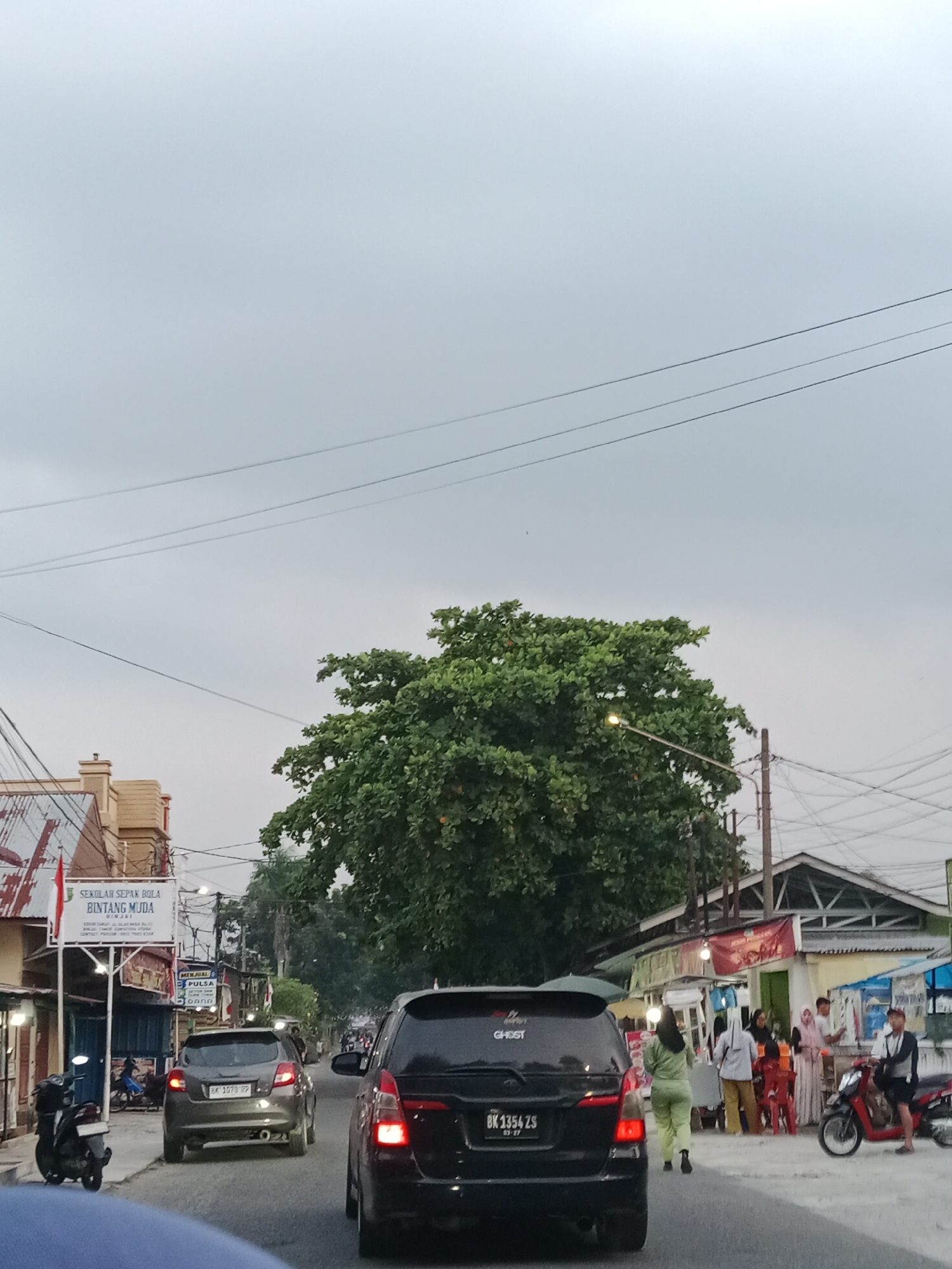 The image captures a street scene in the city of Binjai, Indonesia, likely during the late afternoon or early evening due to the overcast sky and streetlights beginning to illuminate. The perspective is from inside a vehicle, looking forward and slightly to the right.
In the foreground, the rear of a black SUV with a license plate reading "BK 1354 ZS" is visible. To its left, a darker vehicle is slightly out of focus. The road is paved and appears to be a two-lane street with some traffic.
On the left side of the road, a building with a sign that reads "SEKOLAH SEPAK BOLA BINTANG MUDA" (Star Young Football School) is prominent. Next to it, a scooter is parked. Further down the street, there are more shops and stalls, with people milling about. One stall has a sign that says "MENJUAL PULSA" (Selling Credit).
A large, lush green tree dominates the mid-ground, partially obscuring some of the buildings and a streetlamp that is lit. The streetlamp is positioned to the right of the tree.
On the right side of the street, there are more stalls and shops. People are gathered around these stalls, some appearing to be customers and vendors. A person wearing a light green outfit is walking away from the camera. A motorcycle with a rider is parked near one of the stalls.
The overall environment suggests a busy urban street with commercial activity. The sky is a pale gray, indicating diffused light. Power lines are visible crisscrossing the sky, typical of urban settings in Indonesia.